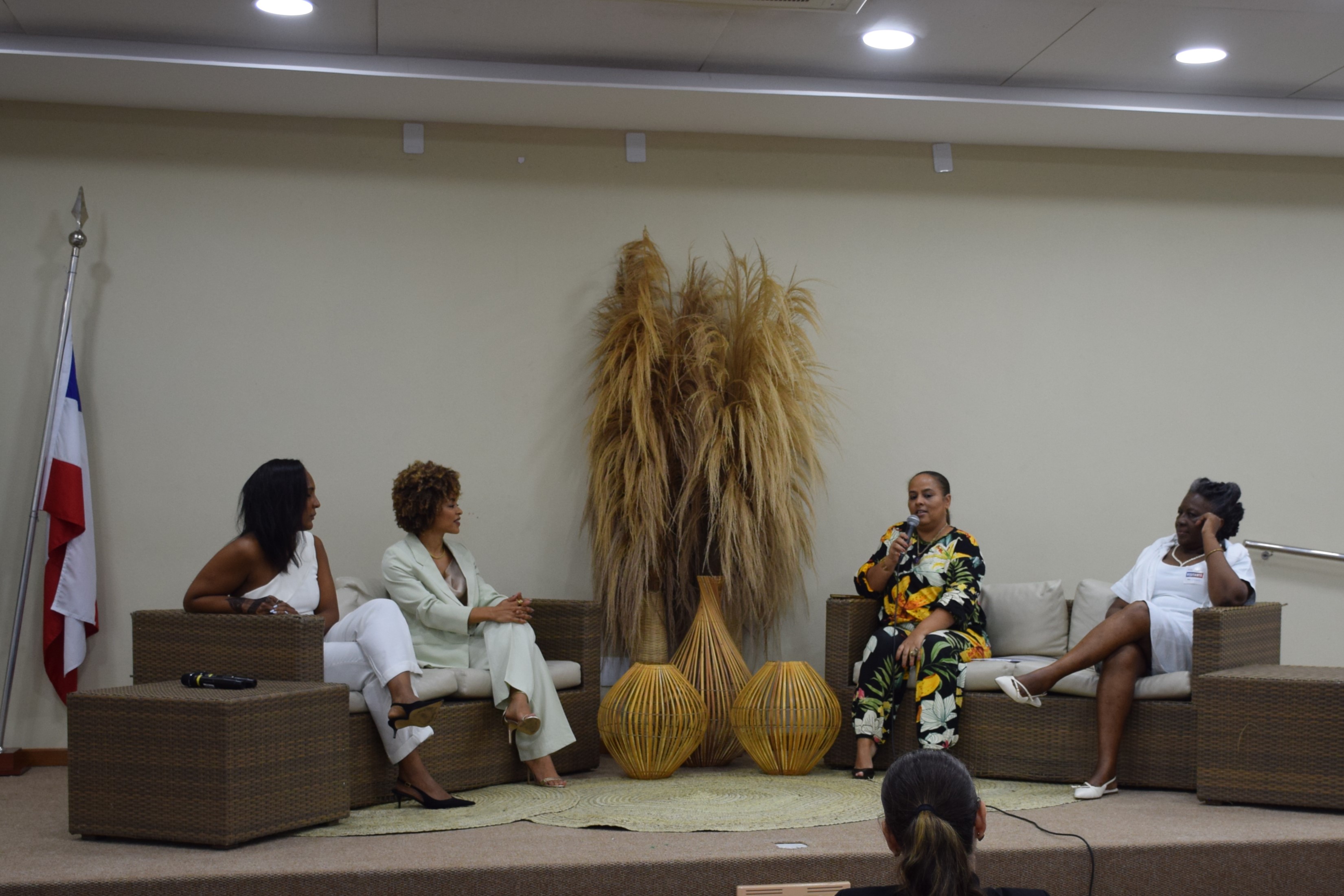 Quatro mulheres negras participam de uma roda de conversa em um auditório. Elas estão sentadas em poltronas de vime. A mulher no centro, com roupa estampada em tons de amarelo, verde e preto, fala ao microfone. As outras três mulheres estão ouvindo atentamente. Ao fundo, há três grandes vasos decorativos e bandeiras do Brasil e da Bahia.