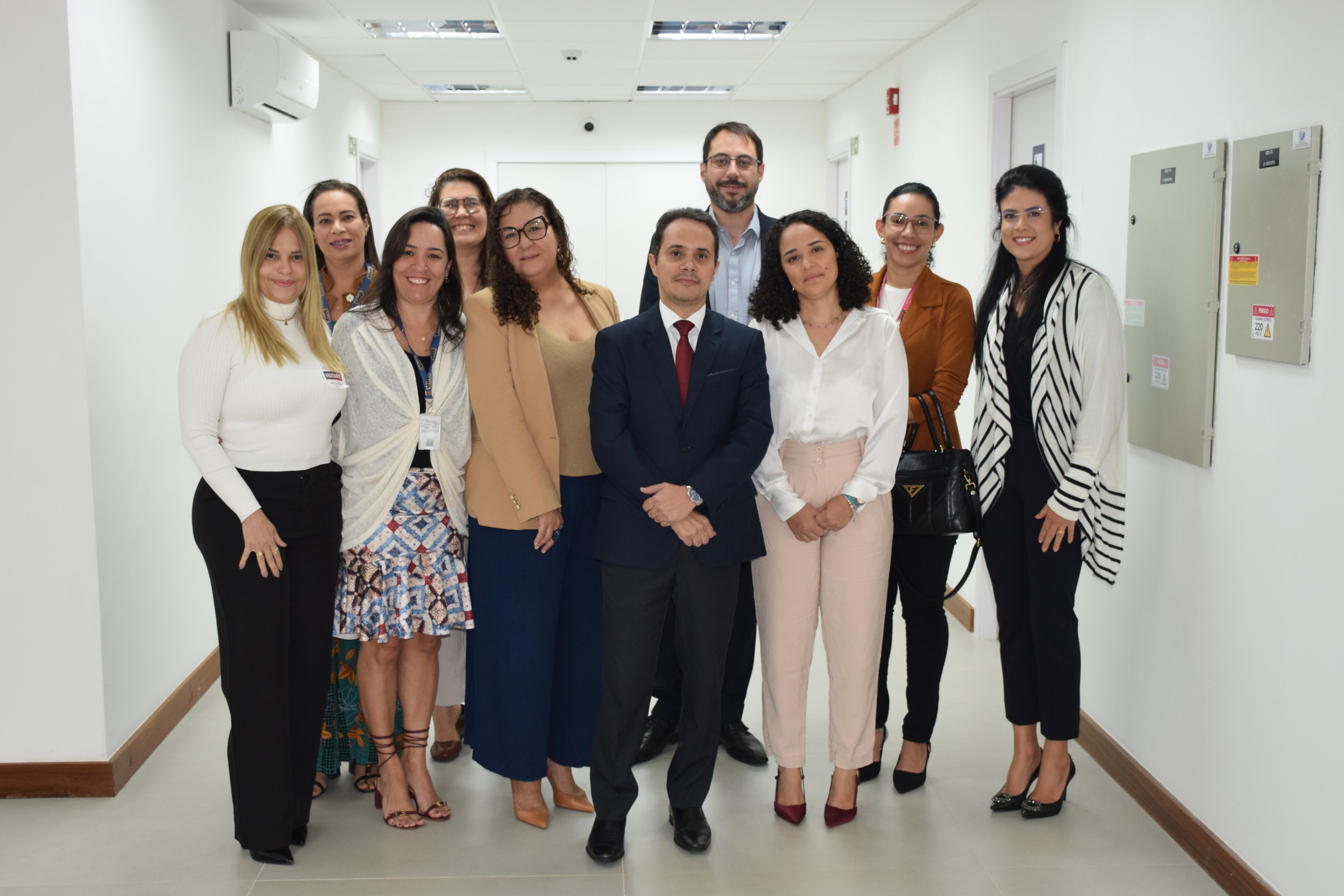 Grupo de 10 pessoas, entre homens e mulheres, posando para foto em um corredor de ambiente corporativo. Todos estão sorrindo e vestidos com trajes formais ou sociais.