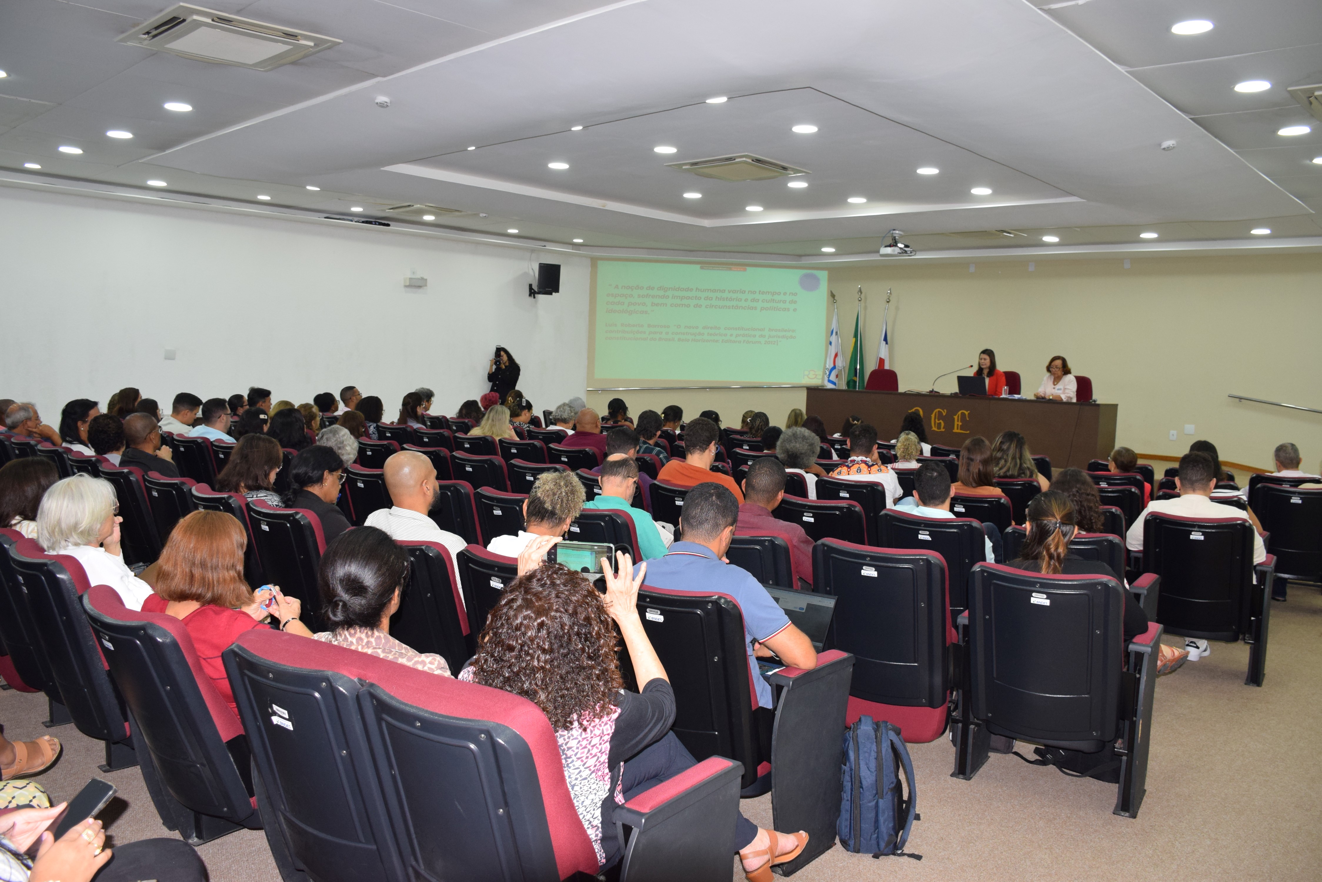Auditório com várias fileiras de cadeiras ocupadas por pessoas assistindo a uma apresentação. Duas mulheres estão sentadas à mesa no palco, uma delas falando ao microfone. Atrás delas, há quatro bandeiras e uma projeção com texto na parede. O ambiente é iluminado com luzes no teto e tem aparência formal e organizada.