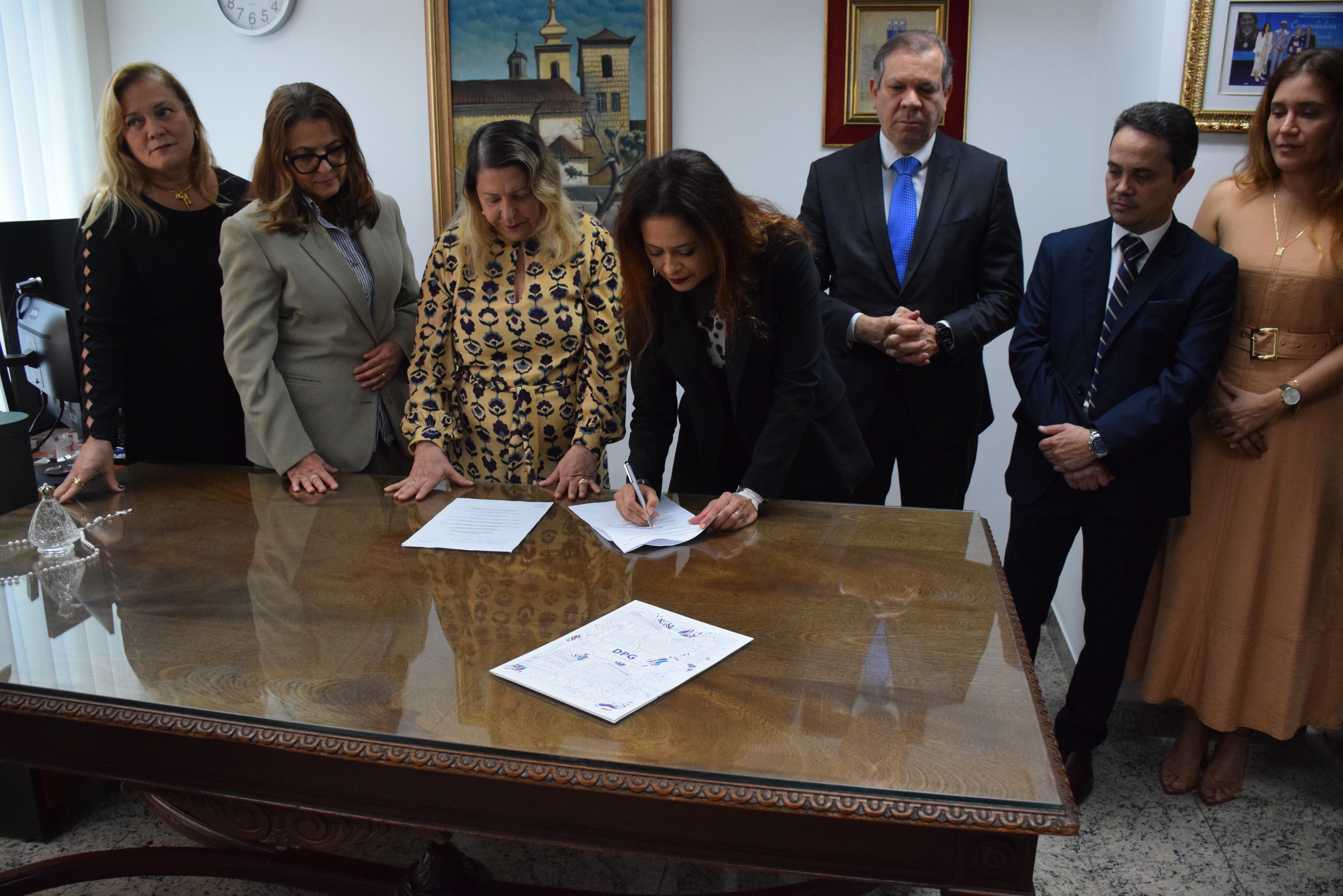 A imagem mostra um grupo de 7 pessoas em um ambiente interno. Uma mulher, posicionada à frente, está assinando um documento sobre a mesa. Ao redor dela, as outras pessoas observam atentamente, algumas com as mãos unidas na frente. As pessoas estão vestidas de forma formal. No fundo, é possível ver quadros nas paredes e um relógio. A mesa é de madeira e contém documentos e uma peça decorativa. O ambiente é bem iluminado.