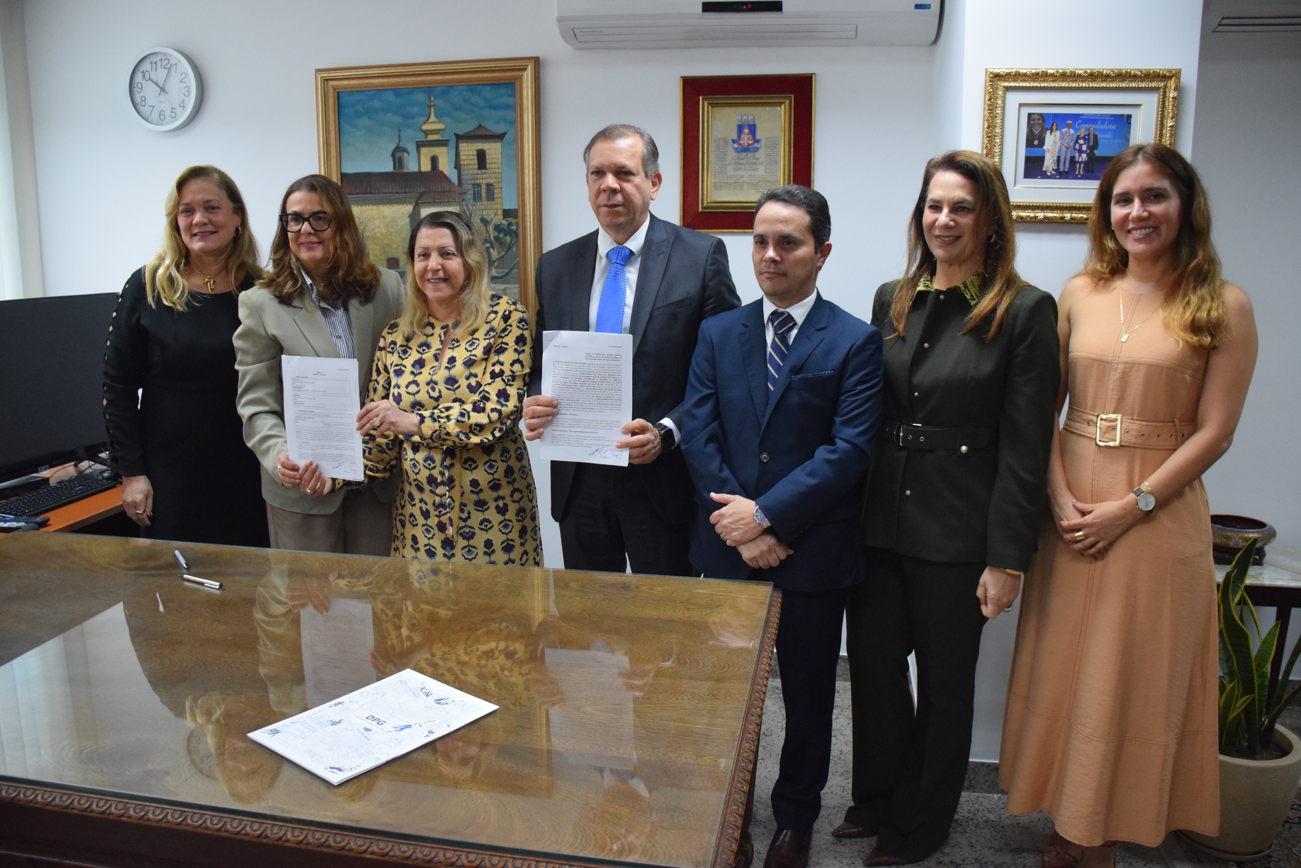 A imagem mostra sete pessoas em um ambiente interno. Elas estão posando para a foto, sorrindo e segurando documentos em suas mãos. O fundo da imagem tem quadros nas paredes, um relógio e uma planta ao lado. A mesa de madeira está coberta com mais documentos. Todos estão vestidos de forma formal, com roupas de cores variadas. O ambiente é bem iluminado.
