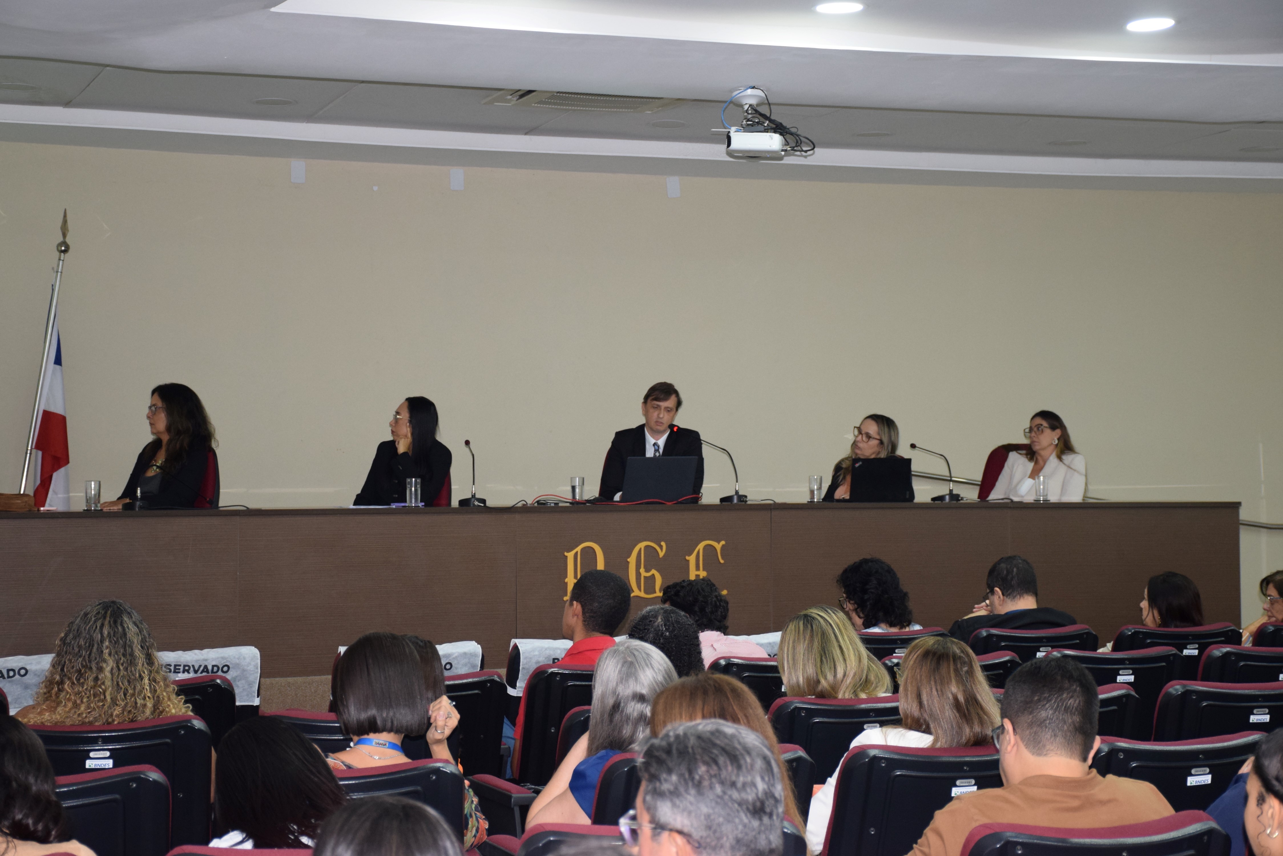 Em uma sala de auditório, várias pessoas estão sentadas assistindo a uma mesa de debate. Ao fundo, cinco pessoas estão sentadas atrás de uma bancada com microfones e laptops. No centro da mesa, um homem de terno fala ao microfone. As outras quatro pessoas, todas mulheres, ouvem atentamente. À esquerda da imagem, há uma bandeira do estado da Bahia. Na parte frontal da bancada está escrita a sigla "PGE". O ambiente é iluminado, e o público está atento à apresentação.