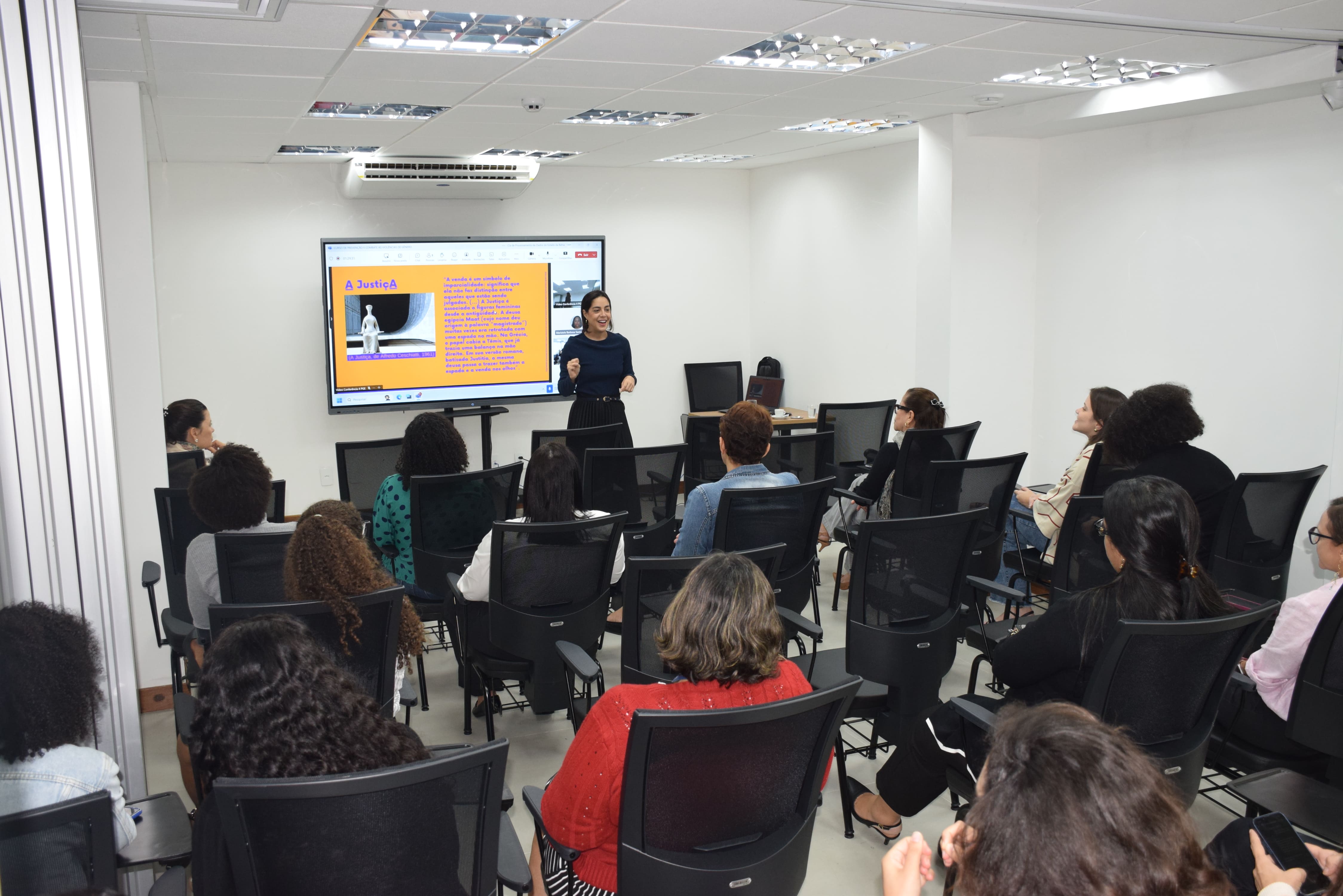 Palestrante apresenta conteúdo sobre Prevenção e Combate em uma sala com várias pessoas sentadas assistindo, olhando para uma tela grande ao fundo com slide colorido.