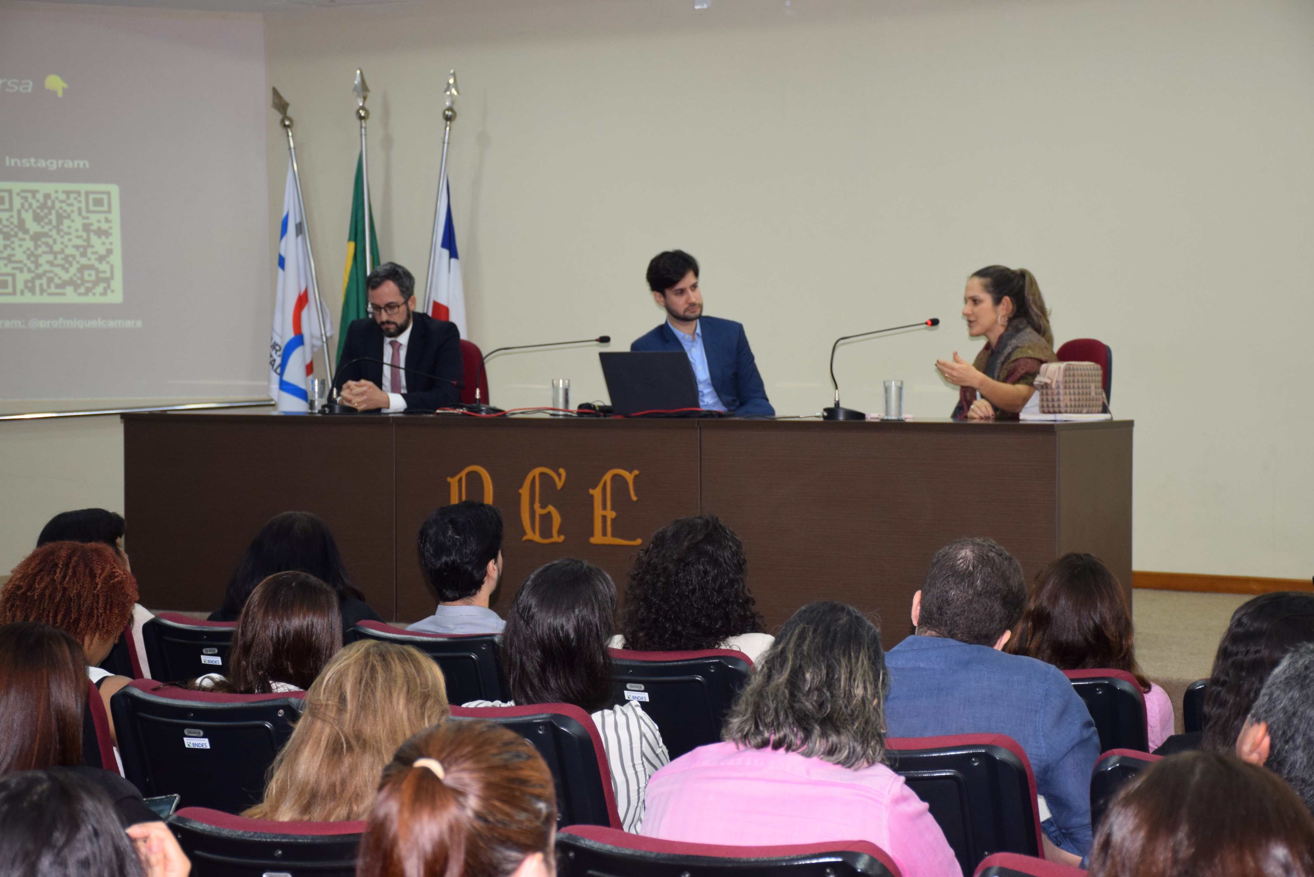 Em um auditório, três pessoas estão sentadas atrás de uma mesa participando de uma mesa-redonda. À esquerda, um homem de terno escuro e gravata vermelha está com as mãos entrelaçadas. No centro, outro homem de terno azul observa a mulher à direita, que fala ao microfone e gesticula com as mãos. Atrás deles, há três bandeiras (Brasil, Bahia e institucional). O público assiste, sentado em cadeiras acolchoadas.