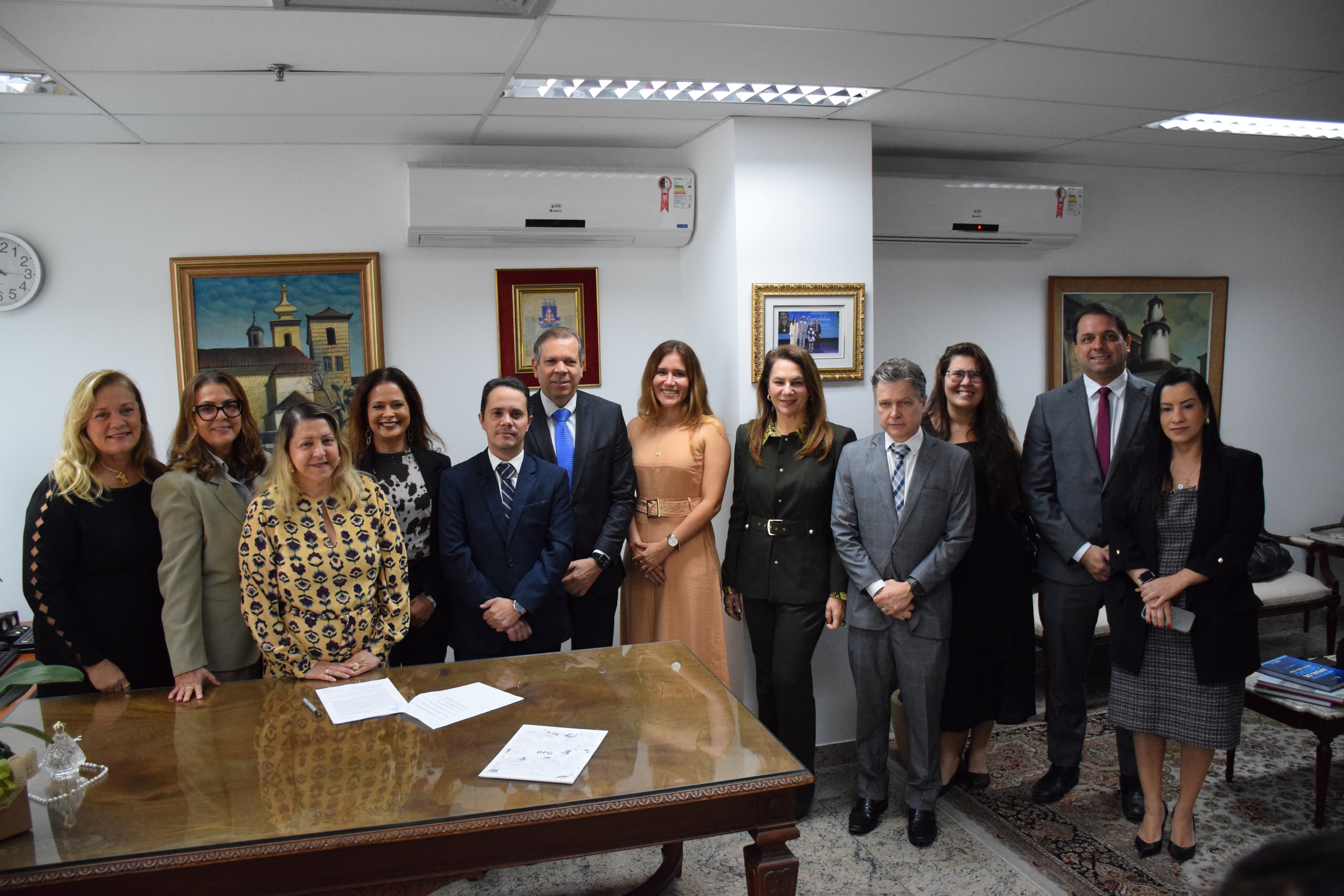 A imagem mostra um grupo de 12 pessoas em um ambiente interno. Eles estão posando para a foto, com 6 pessoas à frente e 6 atrás. As pessoas na frente estão em pé e algumas estão levemente curvadas para a frente, sorrindo. Ao fundo, há quadros nas paredes, e a mesa à frente delas tem documentos e objetos. Todos estão vestidos de forma formal.