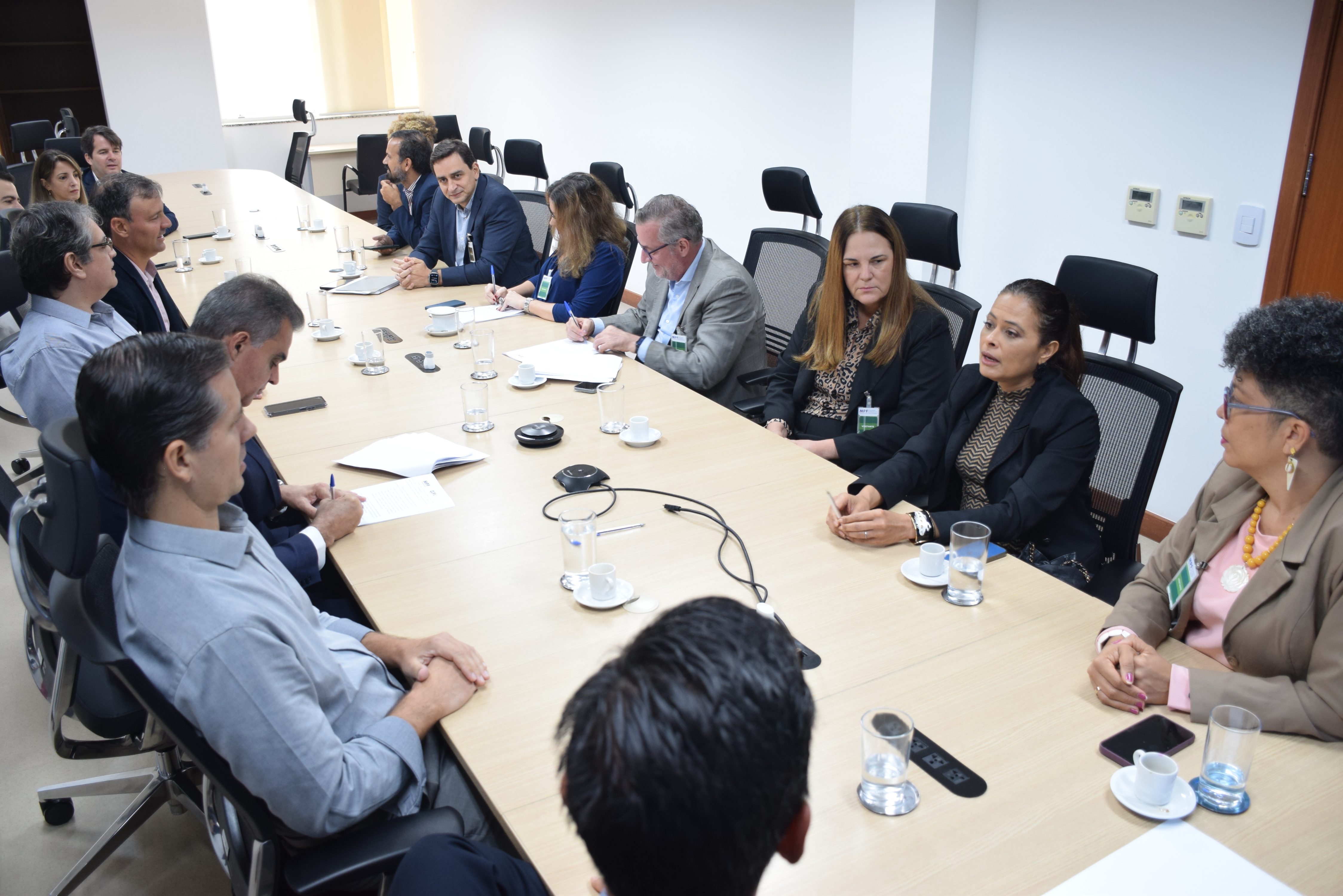A imagem mostra uma reunião em uma sala de conferências. Várias pessoas estão sentadas ao redor de uma mesa retangular. À esquerda, há homens e mulheres, com algumas pessoas escrevendo. À direita, uma mulher de cabelo preso está falando.