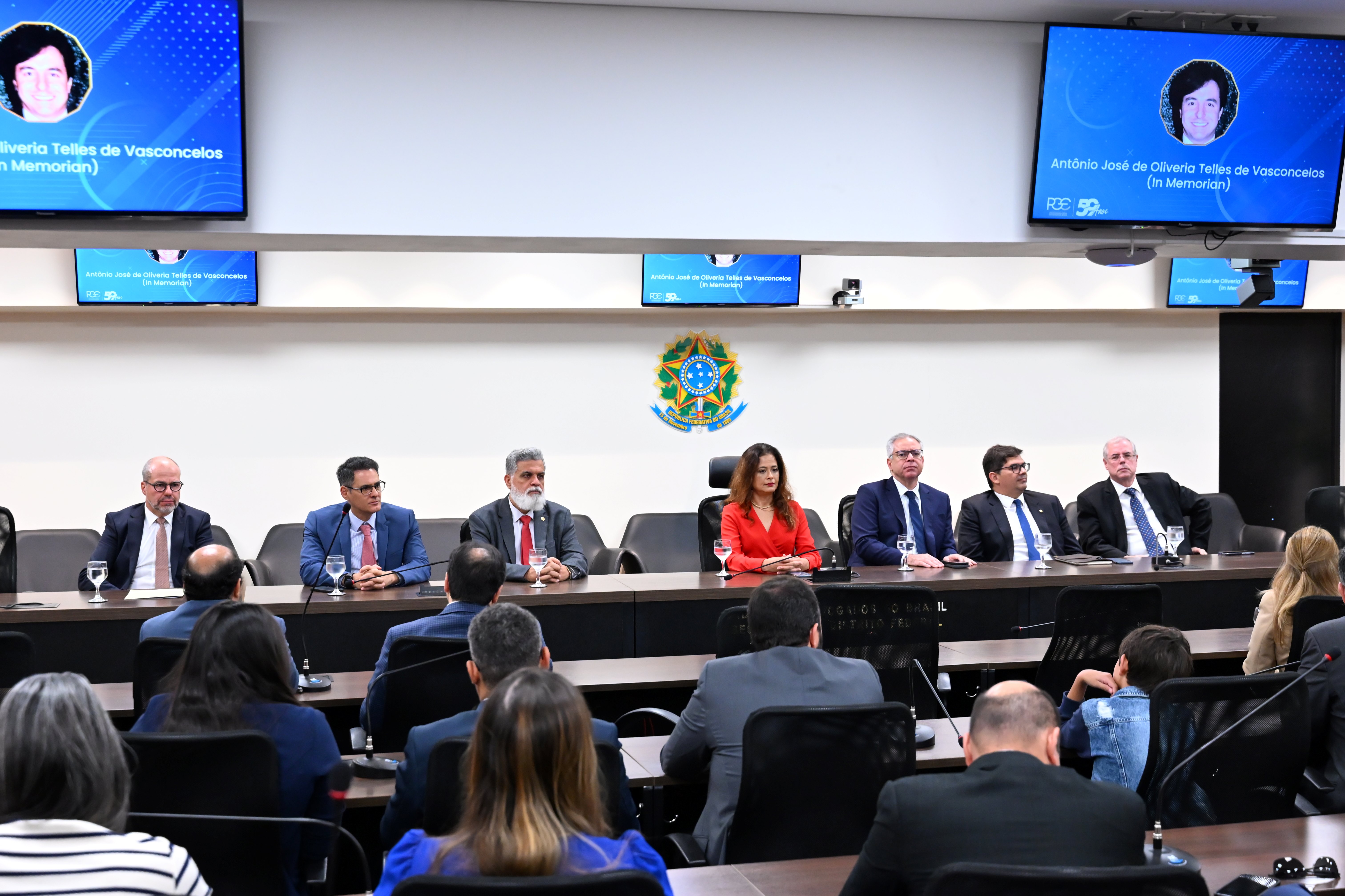 A imagem mostra uma mesa de autoridades em um auditório. Sete pessoas estão sentadas atrás da mesa, sendo seis homens e uma mulher ao centro, vestida com um blazer vermelho. Todos usam trajes formais. À frente, há várias pessoas sentadas assistindo. Na parede ao fundo, há o brasão da República do Brasil e três telas que exibem a foto de um homem com os dizeres: “Antônio José de Oliveira Telles de Vasconcelos (In Memoriam)”. O ambiente é iluminado e transmite formalidade, indicando uma cerimônia de homenagem