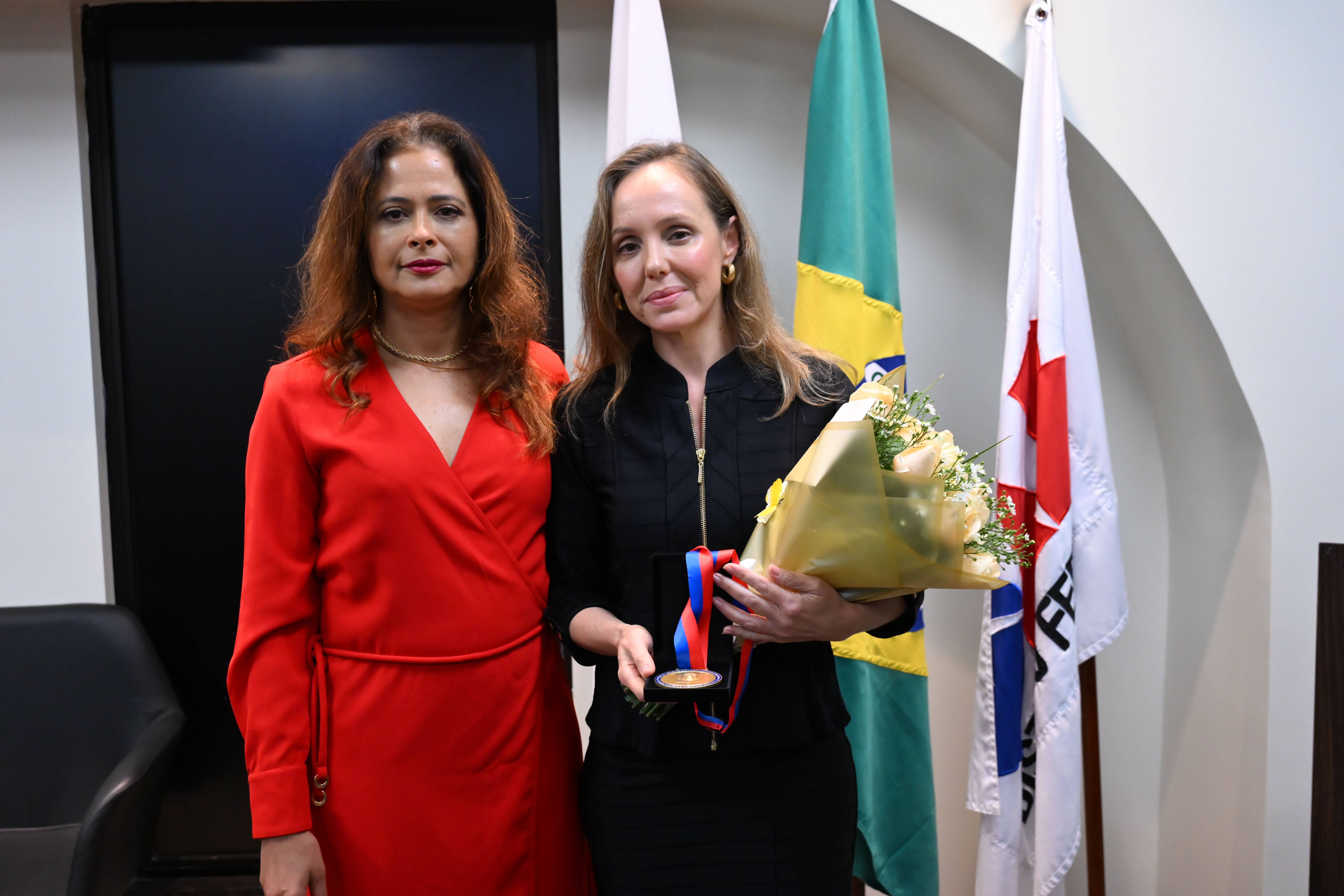 A imagem mostra duas mulheres em pé, lado a lado, olhando para a câmera. A mulher à esquerda usa um vestido vermelho e colar dourado. A mulher à direita veste roupa preta, segura um buquê de flores amarelas e uma medalha com fita nas cores azul e vermelha. Ao fundo, há três bandeiras: uma branca, uma do Brasil e uma com detalhes em vermelho e azul. A cena parece ser de uma cerimônia de homenagem, em ambiente interno e formal.