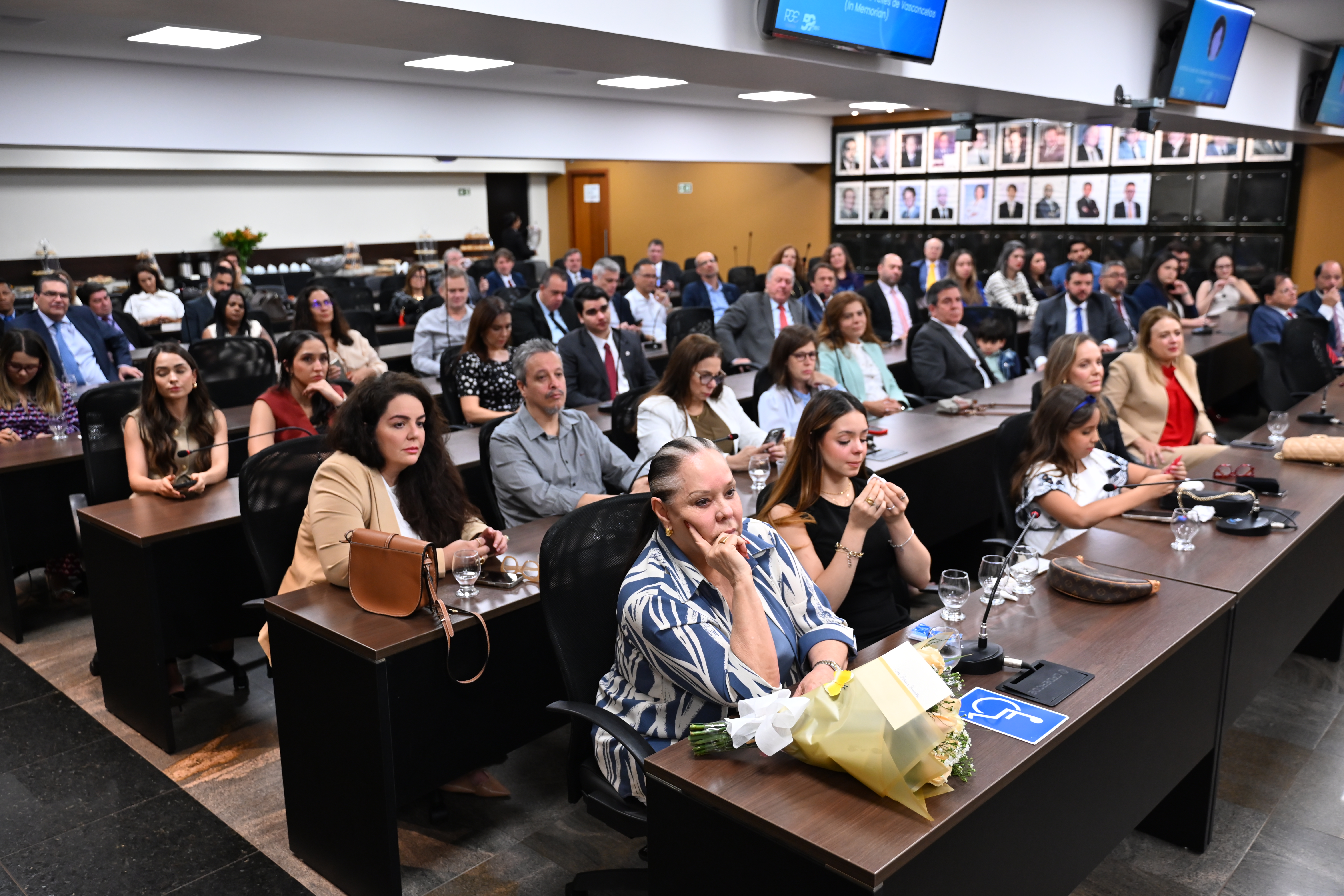 A imagem mostra um auditório cheio de pessoas sentadas em fileiras, assistindo a um evento. Em destaque, na frente, há mulheres e uma criança. As pessoas estão atentas, muitas com expressões sérias. Na parede lateral, quadros com fotos de pessoas. Telas no alto exibem uma homenagem “In Memoriam”. O ambiente é formal e organizado, indicando uma cerimônia.