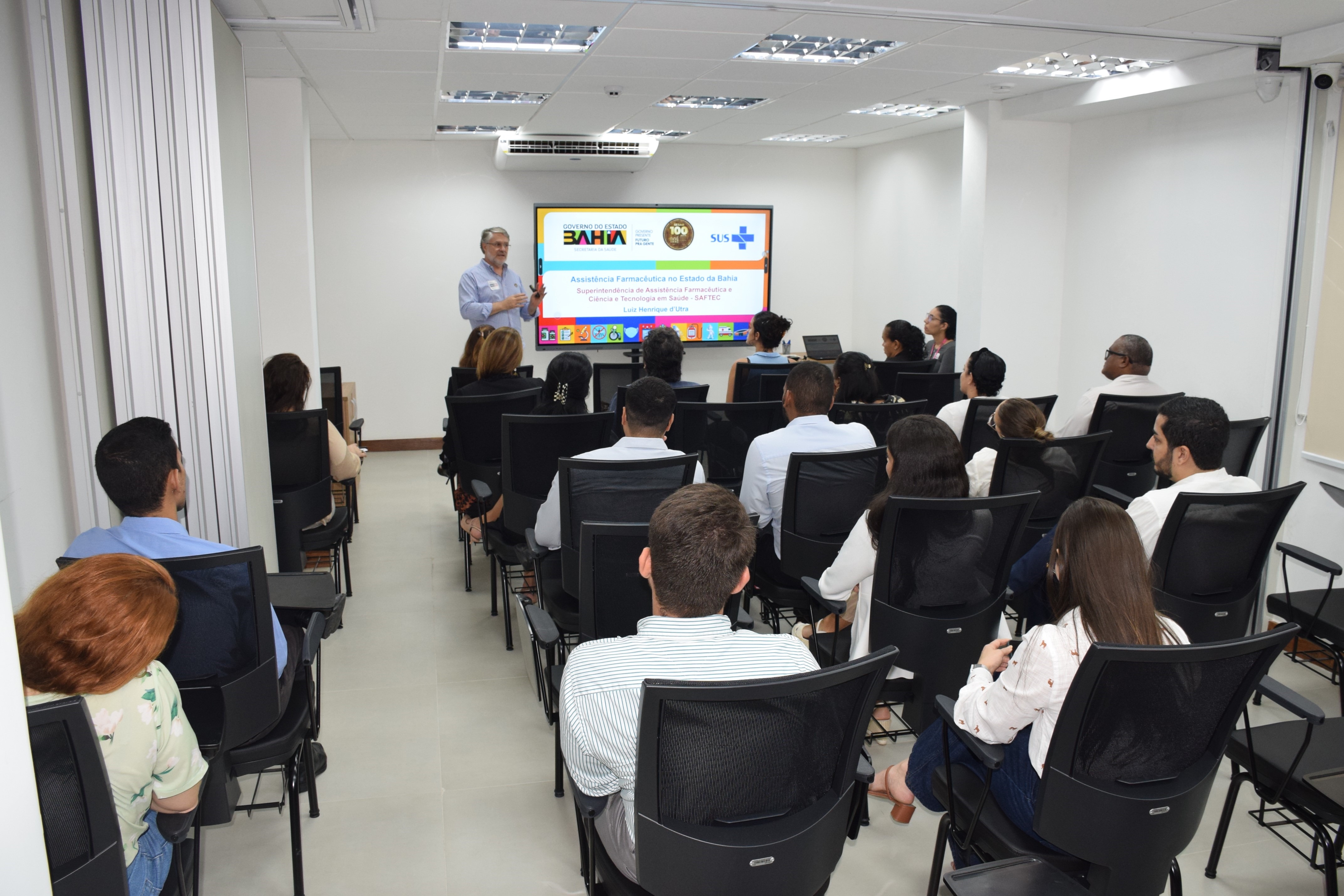 Em uma sala de reuniões clara e moderna, cerca de vinte pessoas estão sentadas, de frente para um palestrante que está em pé ao lado de uma tela. A apresentação exibe o tema “Assistência Farmacêutica no Estado da Bahia”, com logos do Governo da Bahia e do SUS. O público está atento, e a maioria veste roupas sociais. As cadeiras são pretas e a iluminação é branca, vinda do teto. A atmosfera é profissional e de aprendizado.
