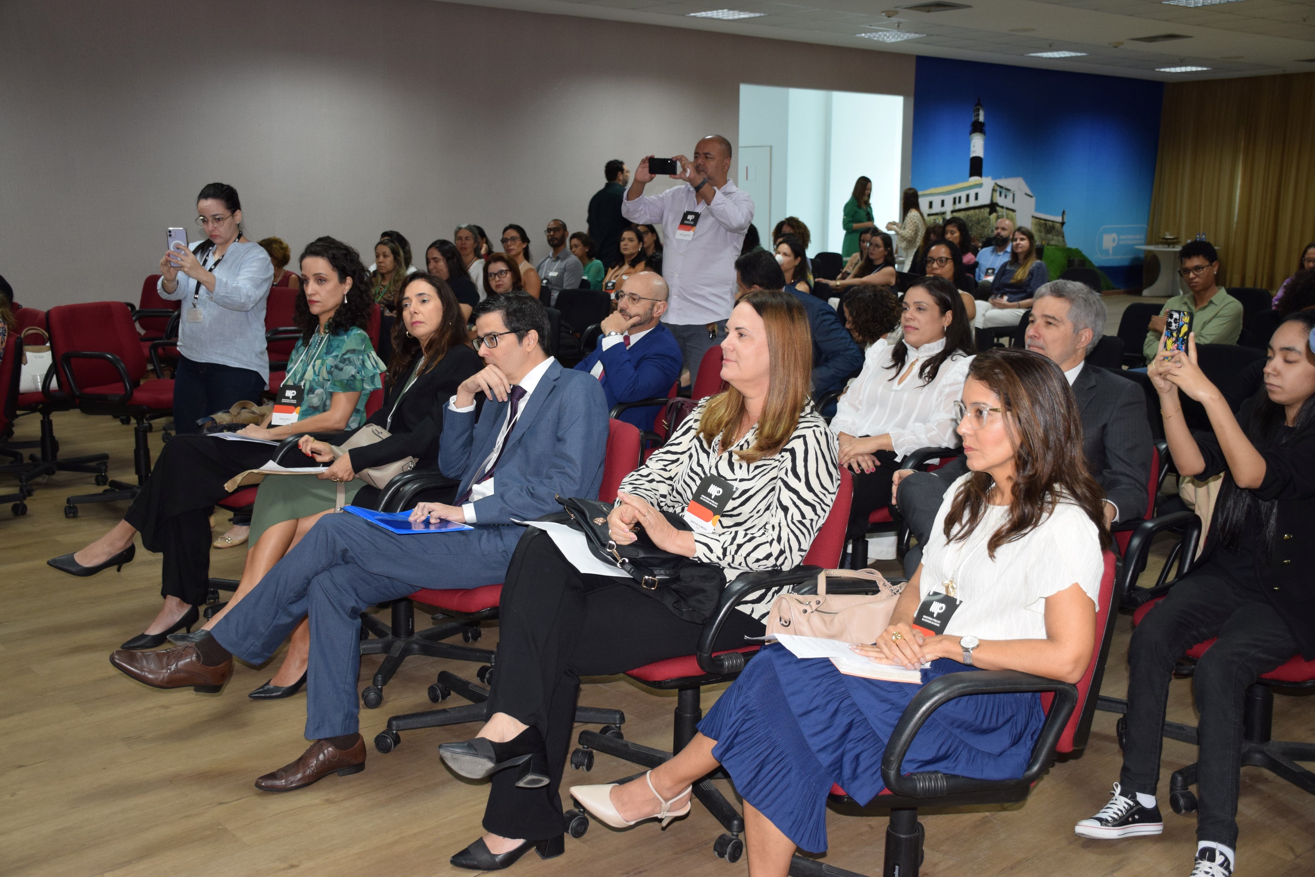 A imagem mostra uma plateia atenta em um auditório. As pessoas estão sentadas em cadeiras vermelhas com rodinhas, e a maioria usa roupas formais. Algumas estão anotando, outras assistindo e algumas tirando fotos com celulares. O ambiente é iluminado, com piso de madeira clara e uma grande imagem do Farol da Barra ao fundo. A cena transmite um clima de evento institucional ou reunião profissional.