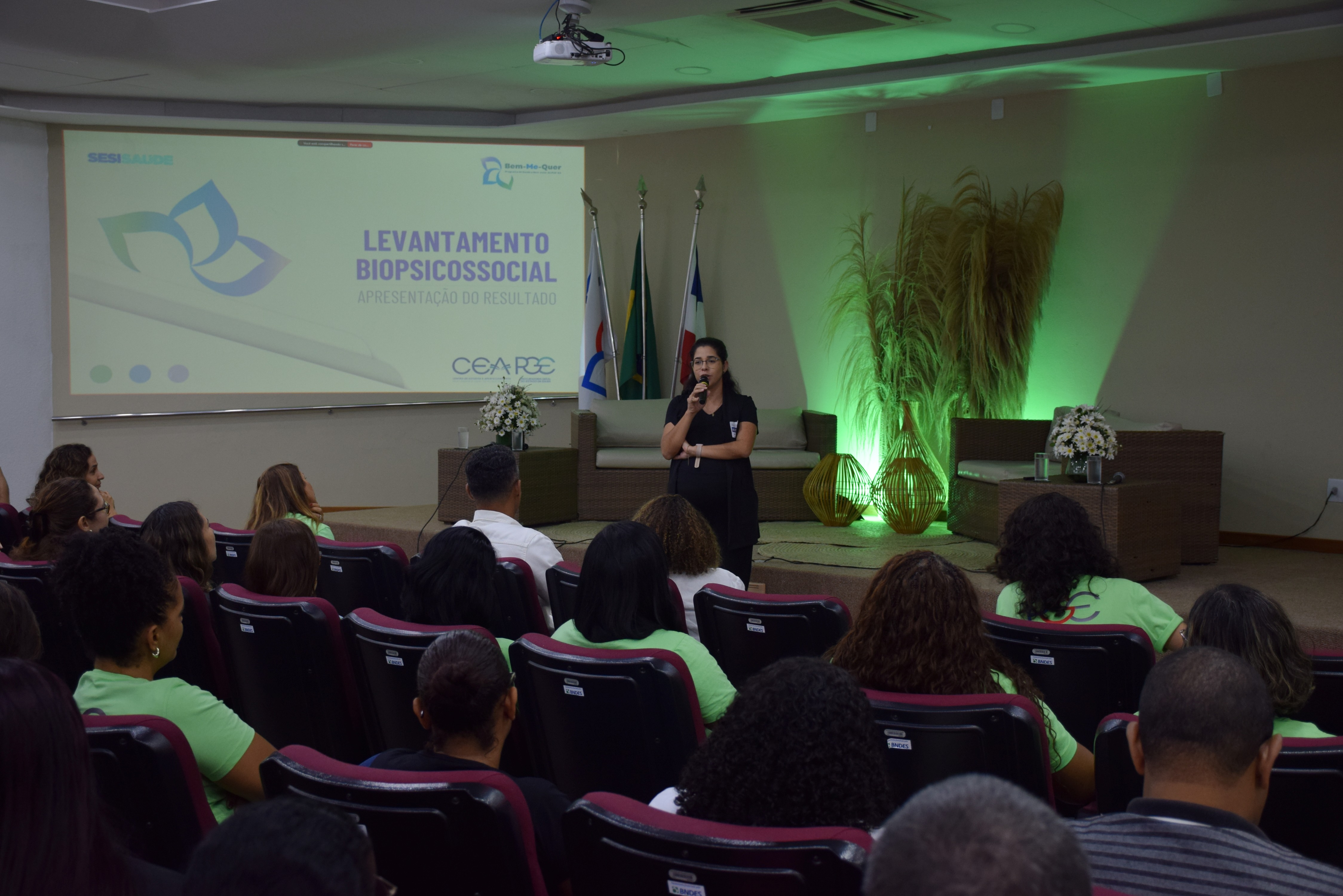 Auditório com público assistindo a uma mulher que está de pé no palco, falando ao microfone. Ela veste roupa preta e está posicionada entre sofás e arranjos decorativos com iluminação verde. Ao fundo, há três bandeiras e um telão exibindo o título da apresentação: “Levantamento Biopsicossocial – Apresentação do Resultado”.