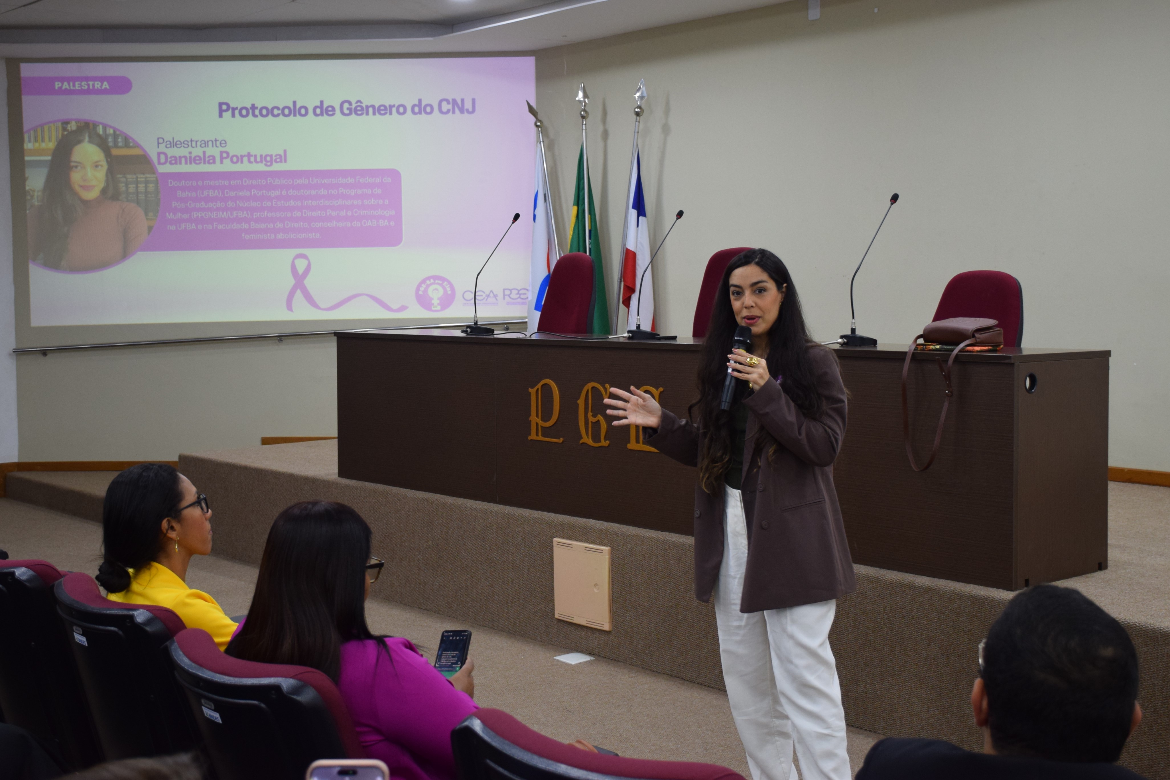 A imagem mostra uma mulher de cabelos longos falando ao microfone em auditório. Ela veste blazer marrom e calça branca. Ao fundo, no telão, aparece a apresentação intitulada “Protocolo de Gênero do CNJ”, com foto e informações da palestrante Daniela Portugal, doutora e mestre em Direito Público pela UFBA. Algumas pessoas sentadas nas poltronas vermelhas acompanham a palestra, enquanto a mesa principal tem bandeiras e microfones.