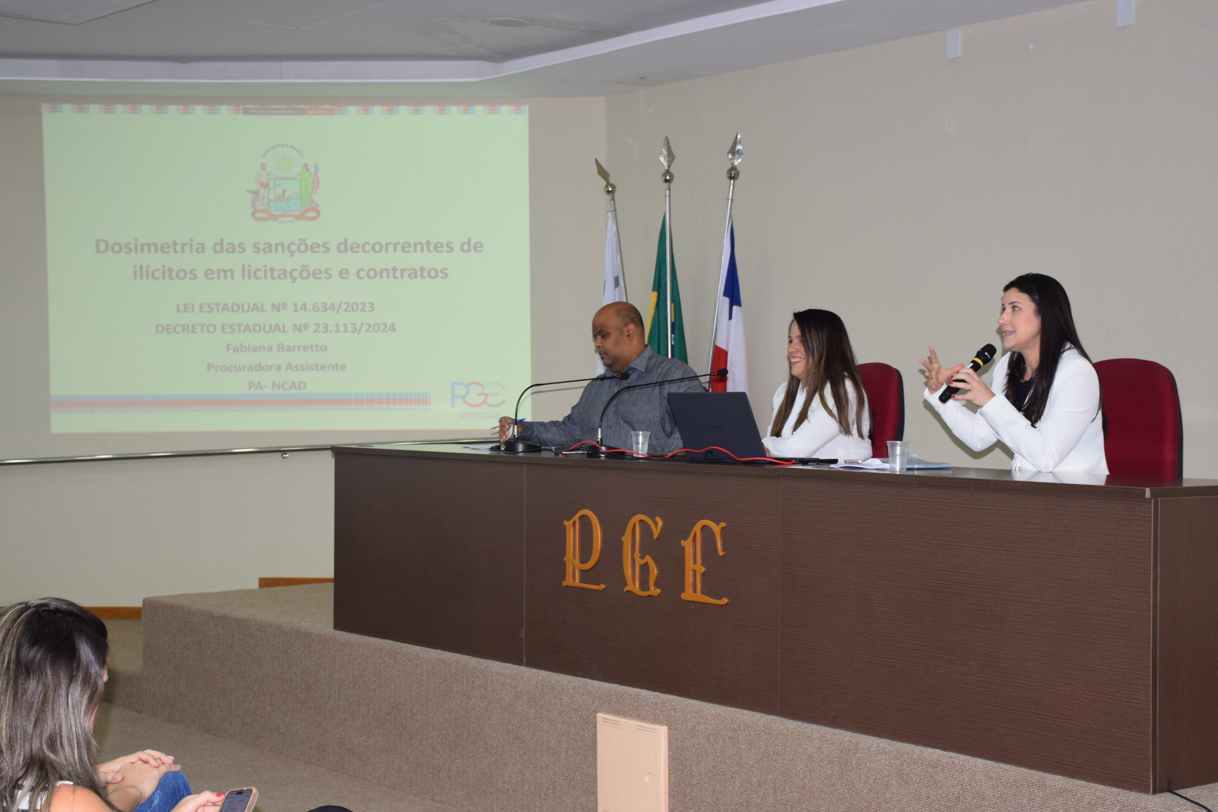 Na imagem, três pessoas estão sentadas atrás de uma mesa de auditório da PGE. À esquerda, um homem de camisa cinza olha para a frente. No centro, uma mulher de blazer branco sorri. À direita, outra mulher de blazer branco fala ao microfone com gestos expressivos. Atrás deles há quatro bandeiras. Na parede, um telão projeta o título da apresentação: “Dosimetria das sanções decorrentes de ilícitos em licitações e contratos”, com referência à lei estadual nº 14.634/2023 e ao decreto nº 23.113/2024.
