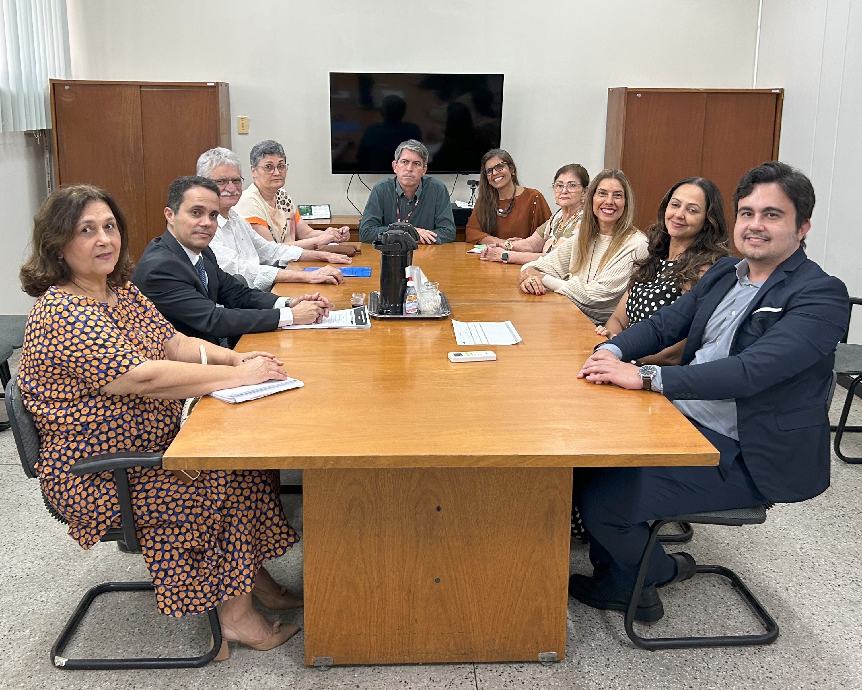 Dez pessoas estão sentadas ao redor de uma mesa retangular de madeira, em uma sala de reunião. Quatro estão à esquerda, cinco à direita e uma ao centro, na cabeceira. Todos olham para a câmera, alguns sorrindo. Sobre a mesa há papéis, canetas, garrafa térmica, copos descartáveis, álcool em gel e controle remoto. Ao fundo, um televisor desligado e dois armários de madeira.