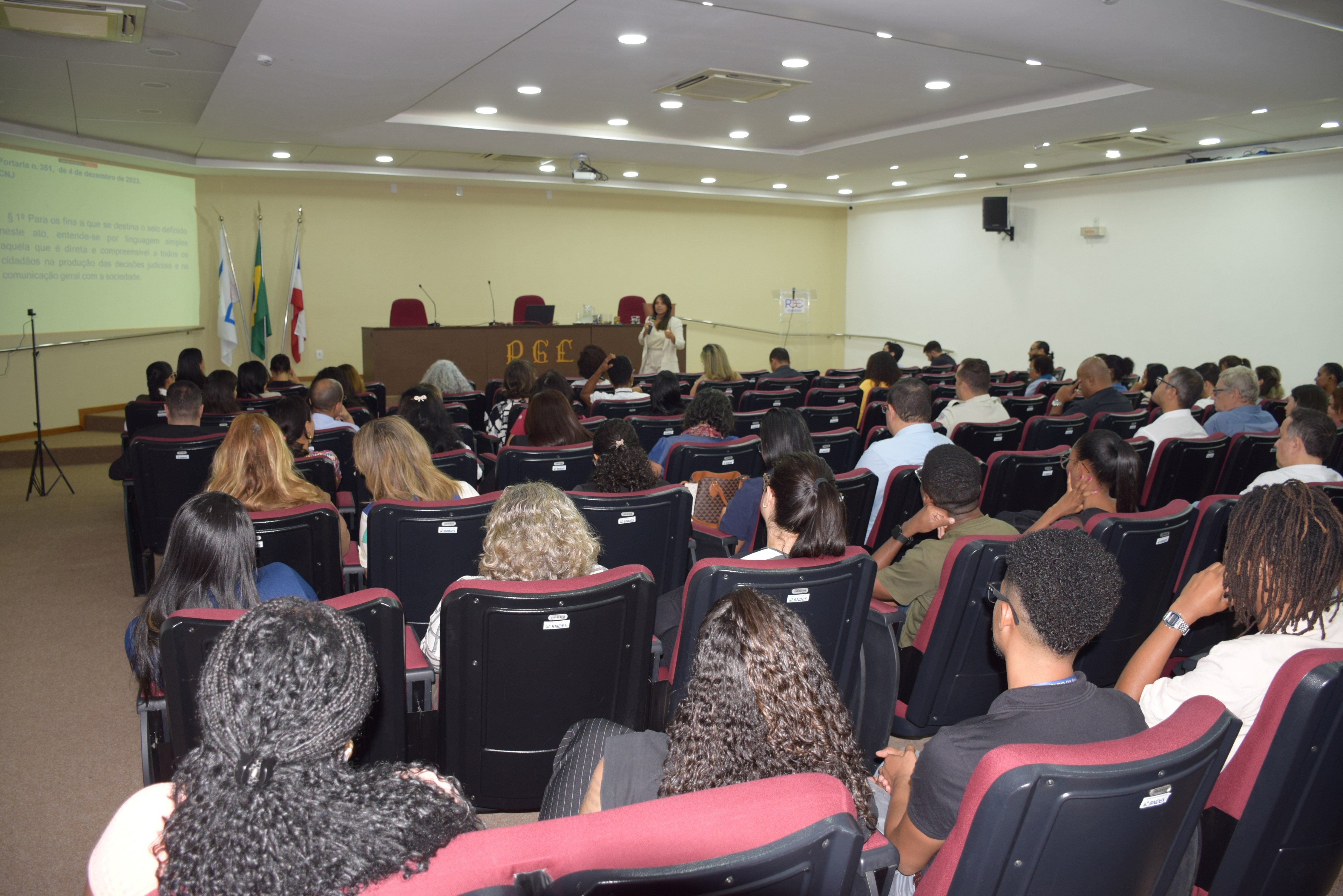 Visão mais ampla do auditório, com a plateia sentada em poltronas vermelhas. A palestrante aparece ao fundo, à frente da tela, explicando o conteúdo jurídico projetado. O local é bem iluminado e há grande participação do público.