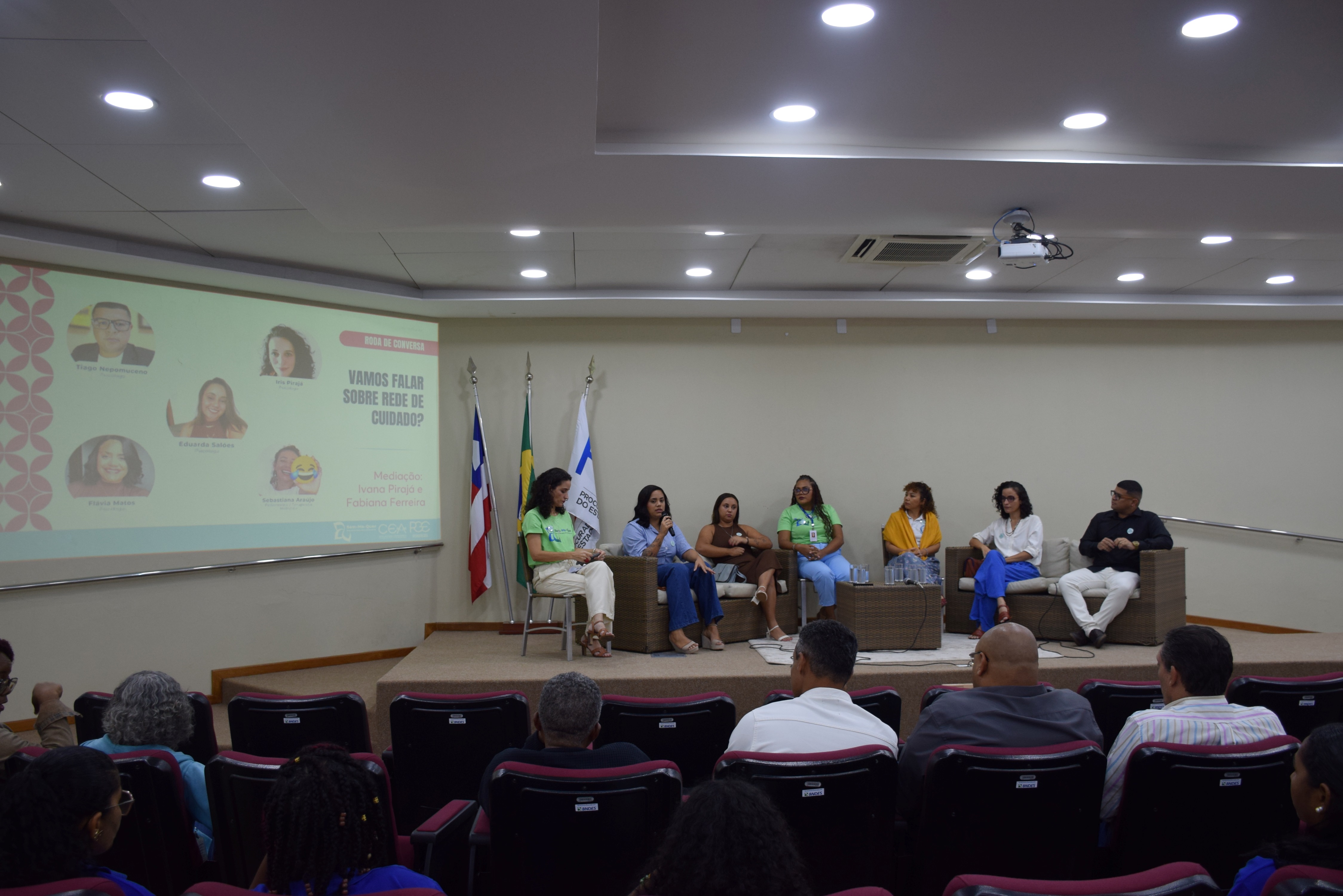 Foto de um auditório com um debate em andamento. No palco, sete pessoas estão sentadas em poltronas de vime, participando de uma roda de conversa. Uma das mulheres fala ao microfone. À esquerda, há um telão com o tema do evento: “Vamos falar sobre rede de cuidado?”. No fundo, há três bandeiras: da Bahia, do Brasil e uma branca institucional. O público está sentado em cadeiras vermelhas, assistindo à conversa. O ambiente é iluminado por luzes de teto.
