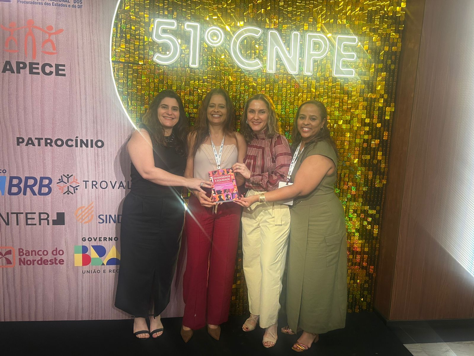 Na imagem, quatro mulheres estão sorrindo e posando juntas em frente a um painel iluminado com a inscrição “51º CNPE”. Elas estão de pé, uma ao lado da outra. A primeira mulher, à esquerda, usa um vestido preto e segura a mão de uma colega. A segunda, de cabelo longo e blusa branca, segura um livro nas mãos. A terceira mulher usa uma blusa vermelha e calça bege. A quarta mulher, à direita, usa um vestido verde e está sorrindo. O fundo é dourado e decorado.