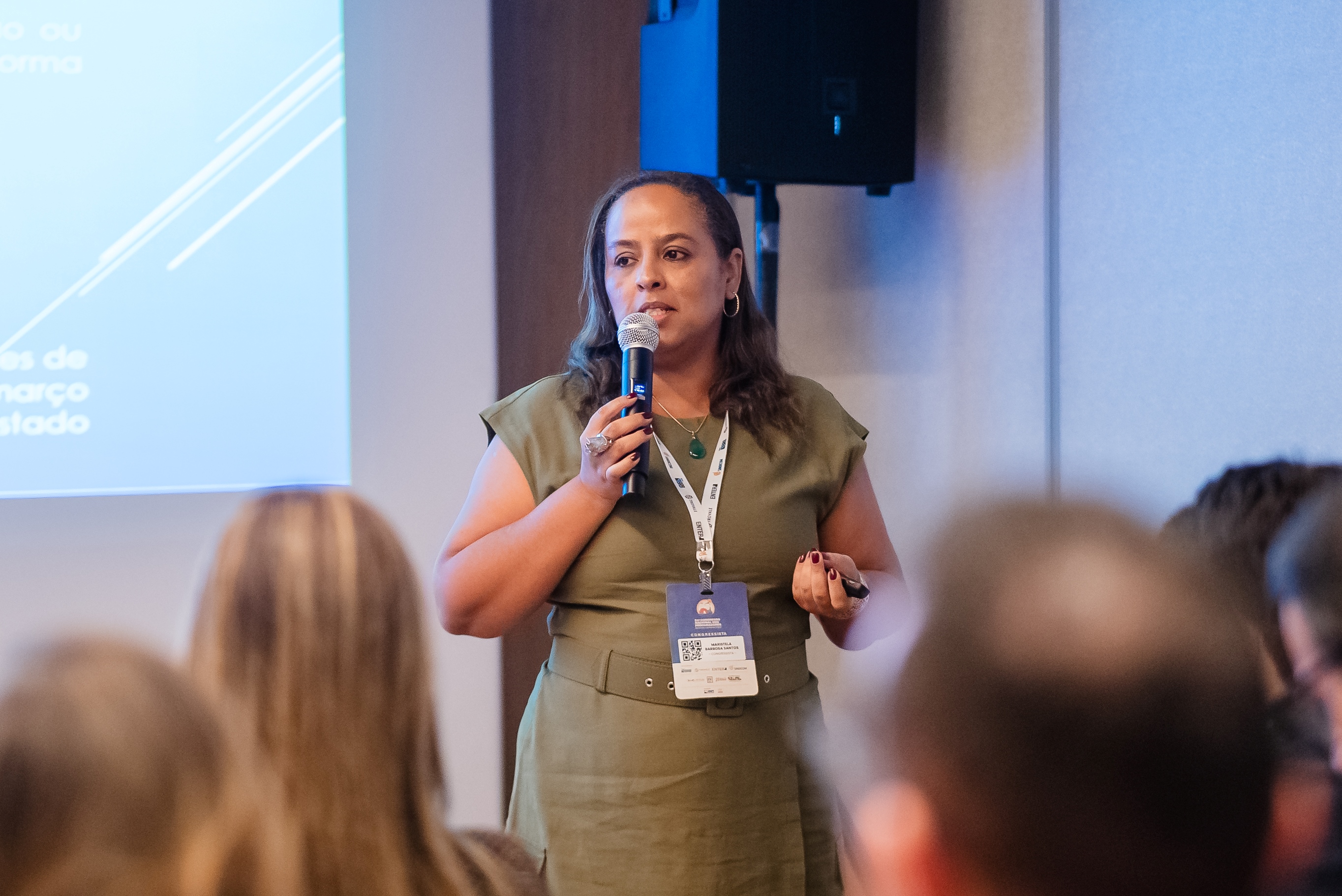 Close da palestrante, mulher de pele clara e cabelos castanhos soltos, usando vestido verde e crachá. Ela segura microfone e controle, enquanto fala ao público atento.