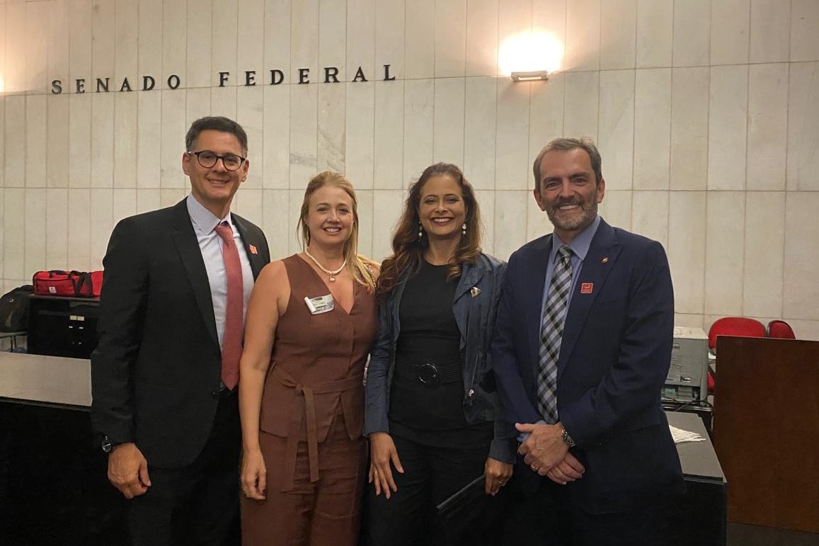 Quatro pessoas posam sorridentes em frente à parede com a inscrição “Senado Federal”. Estão bem-vestidas, demonstrando participação em um evento formal ou institucional. A imagem transmite profissionalismo, união e engajamento em atividades no ambiente legislativo.