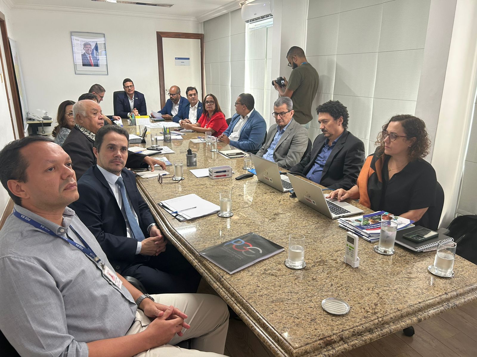 Reunião com 14 pessoas em uma sala de conferência. Homens e mulheres, em sua maioria de terno ou roupas sociais, estão sentados ao redor de uma mesa retangular. Dois notebooks estão abertos. Há copos com água, cadernos e documentos sobre a mesa. O ambiente é iluminado e com paredes brancas.