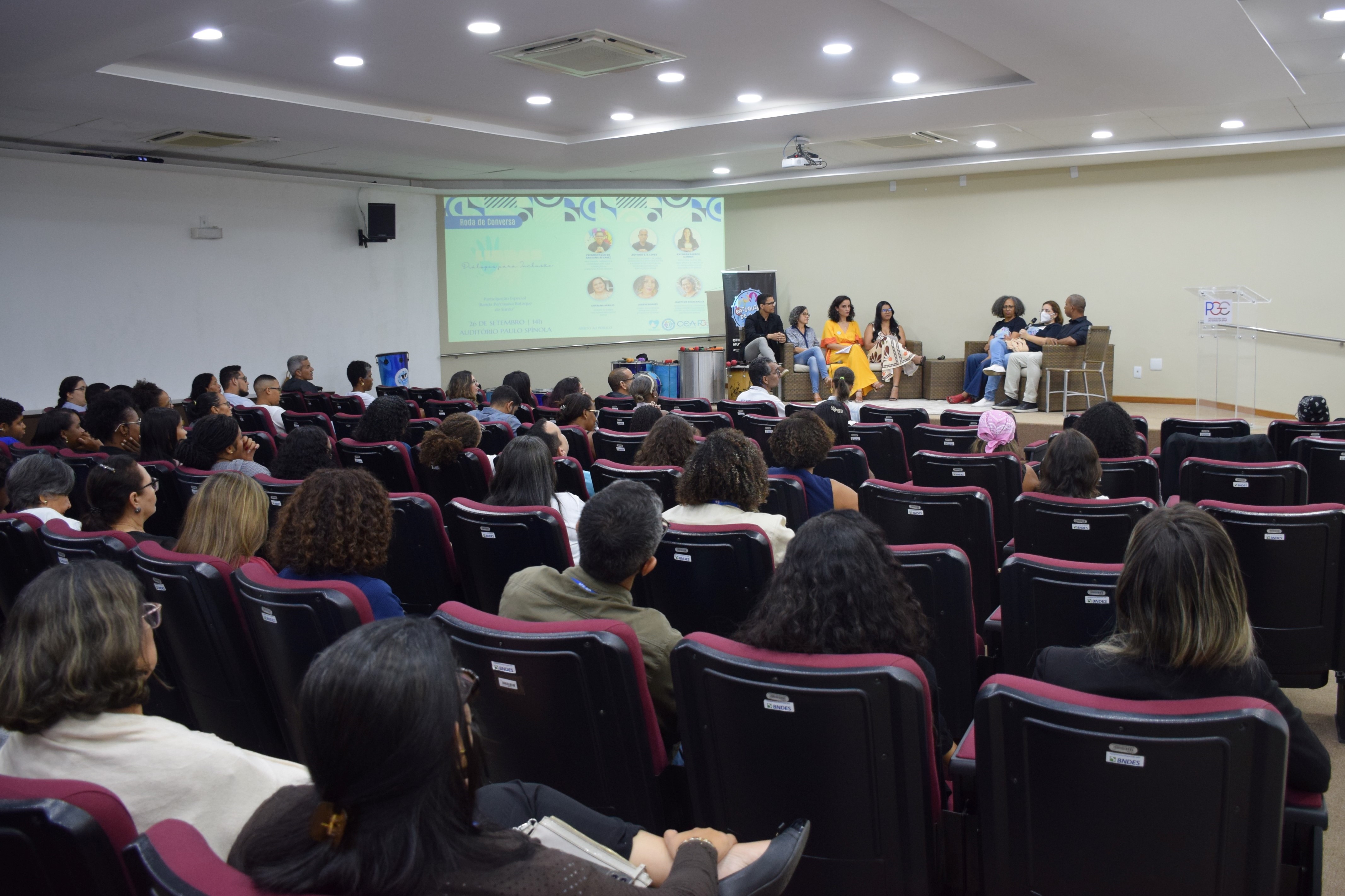 A imagem mostra um auditório com várias pessoas sentadas em cadeiras pretas de encosto alto, voltadas para a frente. No palco, à direita, há um grupo de oito pessoas sentadas em poltronas, participando de uma roda de conversa. Uma tela de projeção ao fundo exibe informações do evento, com fotos e nomes de participantes. À esquerda, o público observa atentamente. À direita, há um púlpito transparente e uma pessoa em pé, interpretando em Libras. O ambiente é iluminado por luzes de teto embutidas.