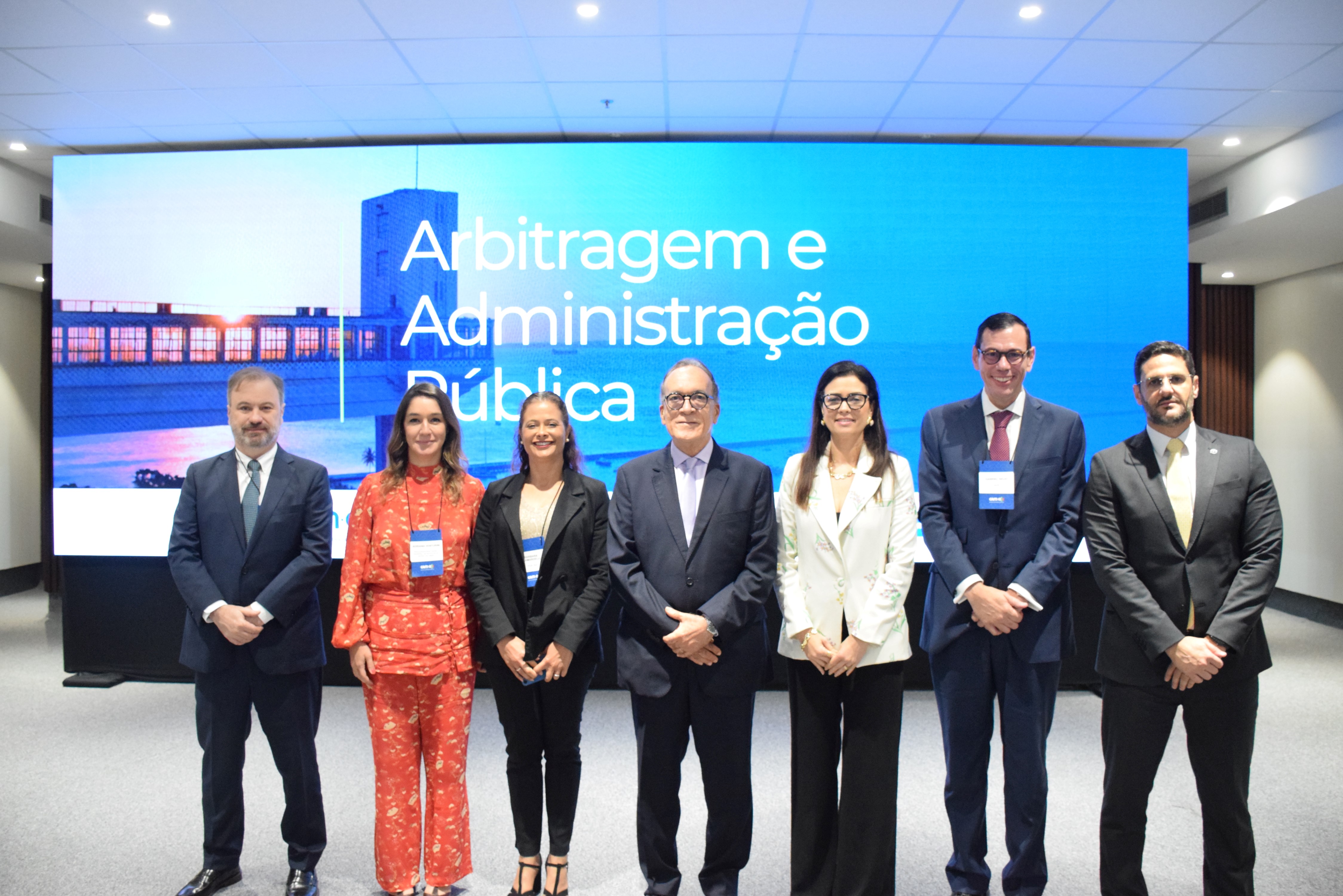 Sete pessoas estão em pé, lado a lado, sorrindo para a foto, em frente a uma grande tela com os dizeres “Arbitragem e Administração Pública”. Quatro homens de terno e gravata e três mulheres bem vestidas, sendo uma com terno floral claro, uma com roupa preta e outra com conjunto vermelho estampado. Todos usam crachá azul pendurado no pescoço. O ambiente é fechado, bem iluminado e com aparência profissional.