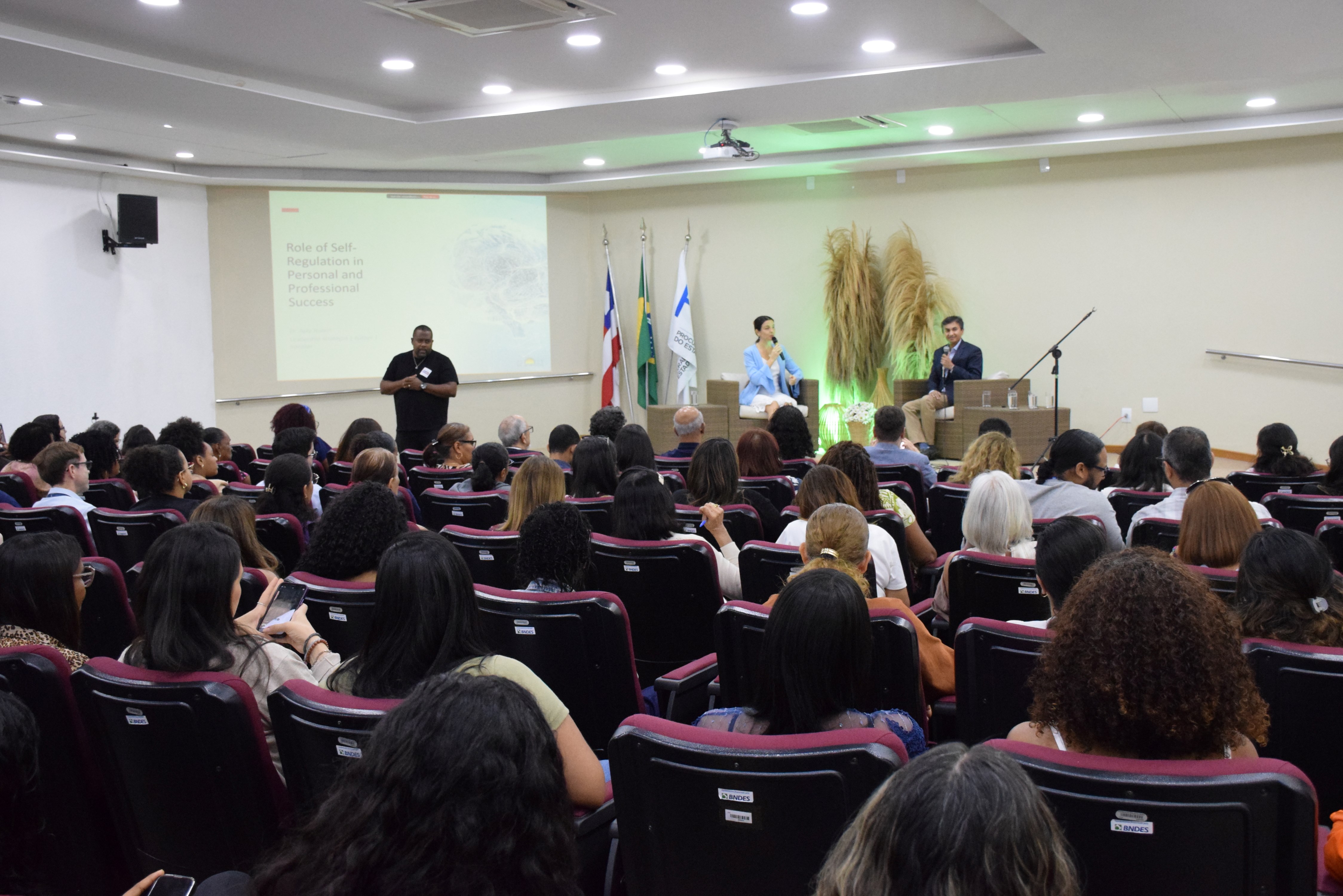 Auditório cheio, com pessoas sentadas em poltronas vinho, voltadas para o palco. No palco, duas pessoas estão sentadas em cadeiras de vime, conversando com microfones, enquanto um homem de preto faz interpretação em Libras no canto esquerdo. Ao fundo, há uma tela com apresentação escrita “Role of Self-Regulation in Personal and Professional Success” e bandeiras da Bahia, do Brasil e de uma instituição. Há também decoração com arranjos de capim seco e iluminação verde suave.