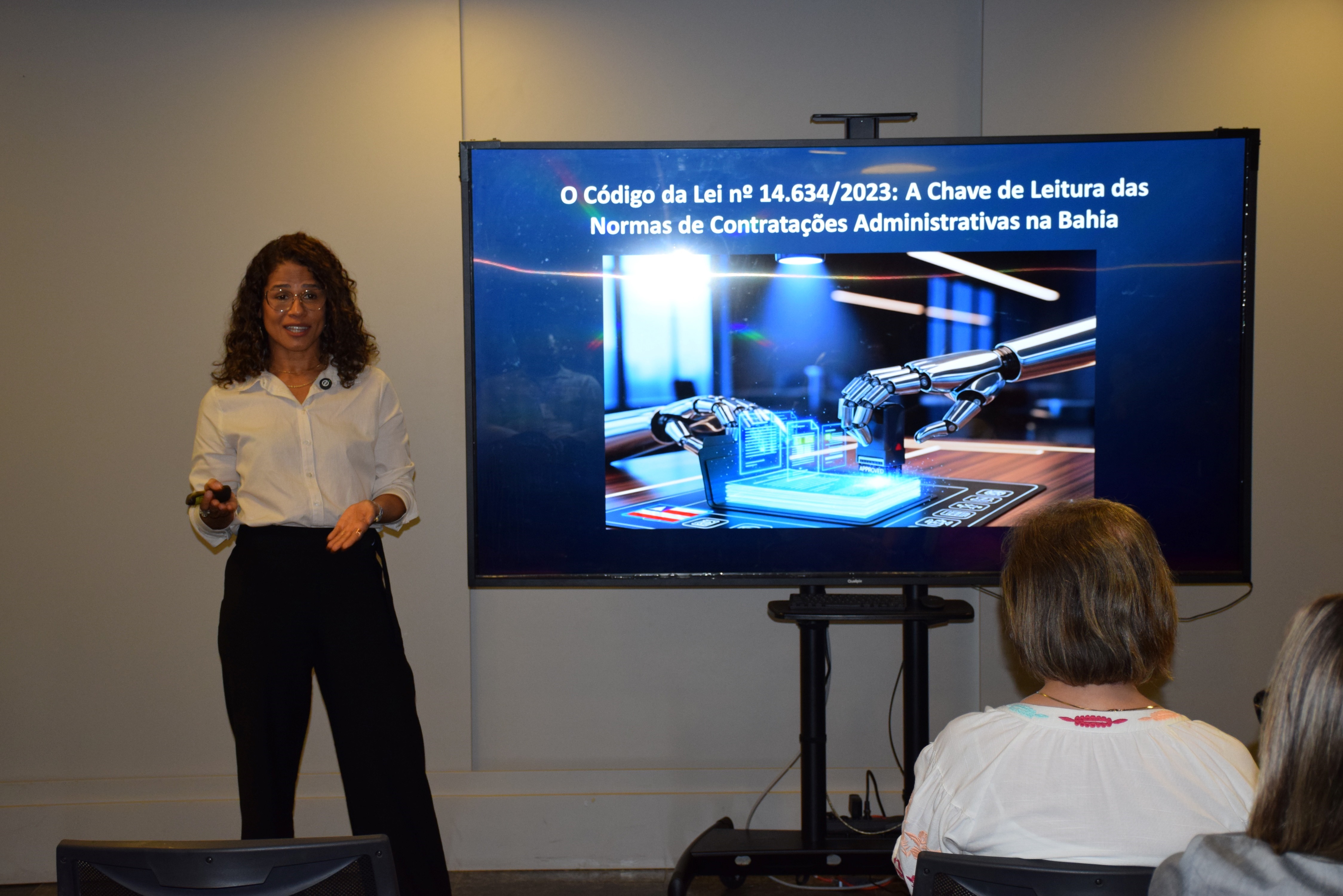 Palestrante apresenta o tema “O Código da Lei nº 14.634/2023: A Chave de Leitura das Normas de Contratações Administrativas na Bahia”. Ela está de pé ao lado de uma tela que exibe uma imagem ilustrativa com braços robóticos manuseando dados digitais, indicando abordagem tecnológica no contexto jurídico. O público acompanha sentado, atento à exposição.