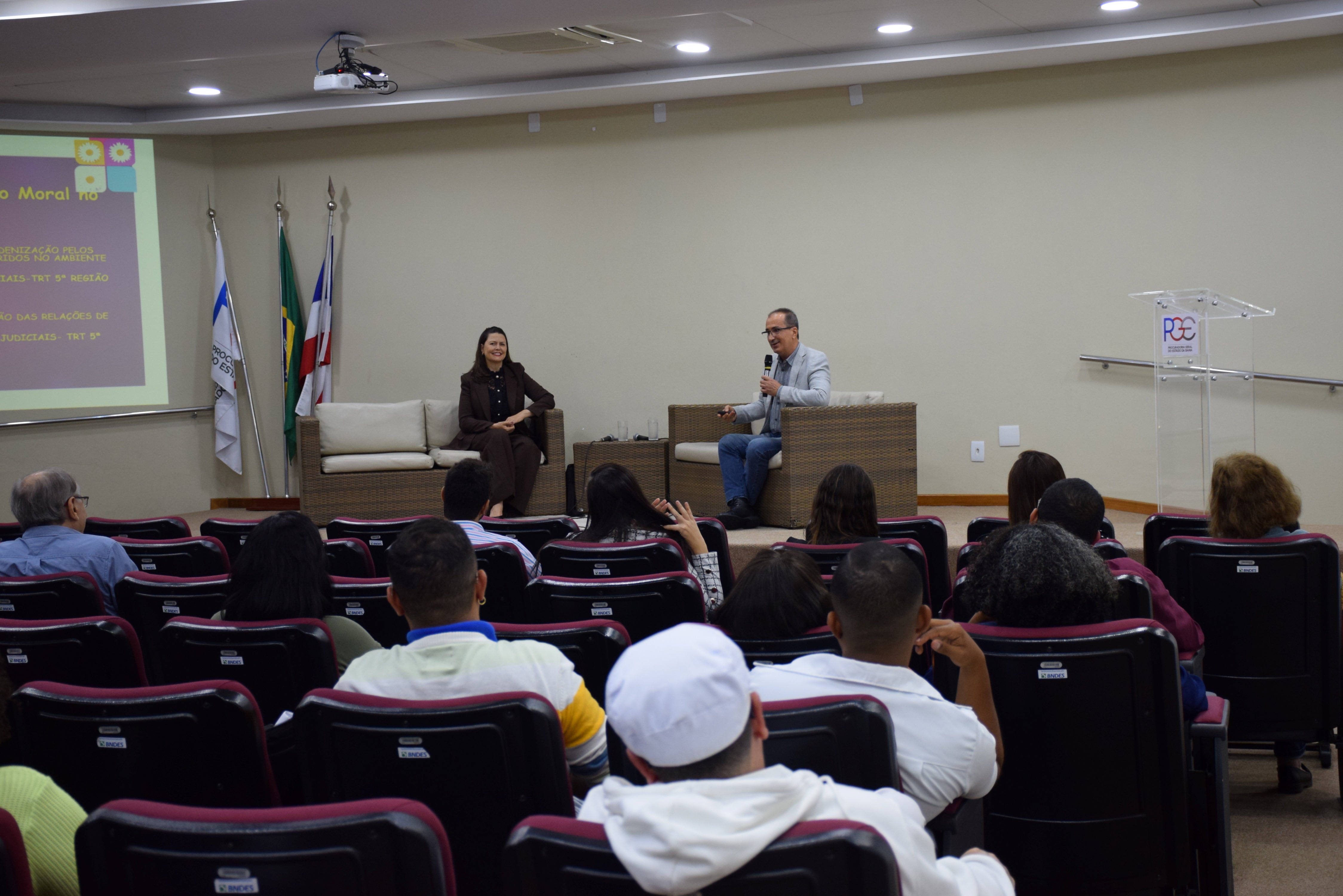 A imagem mostra uma palestra em andamento em um auditório. À frente, um homem fala ao microfone, sentado em uma poltrona de vime com almofadas claras, enquanto uma mulher, também sentada em poltrona semelhante, o observa sorridente. À esquerda, há um telão com apresentação projetada e bandeiras posicionadas ao lado. O público, composto por homens e mulheres, está sentado em cadeiras vermelhas voltadas para o palco. O ambiente é iluminado e organizado.