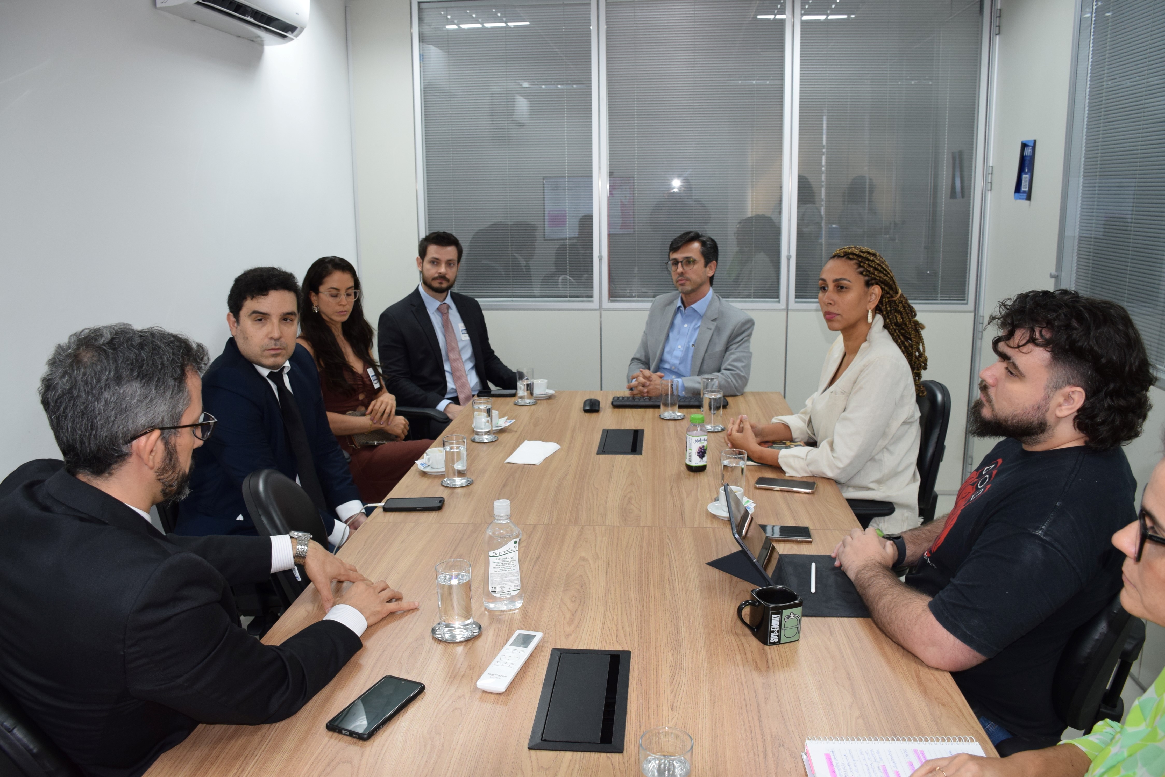 Reunião em uma sala com oito pessoas sentadas ao redor de uma mesa retangular de madeira. Elas estão em conversa atenta, com copos de água, canecas, garrafas e cadernos sobre a mesa. O ambiente é corporativo, com paredes brancas, janelas de vidro e iluminação clara. As pessoas usam roupas formais e demonstram concentração no diálogo.