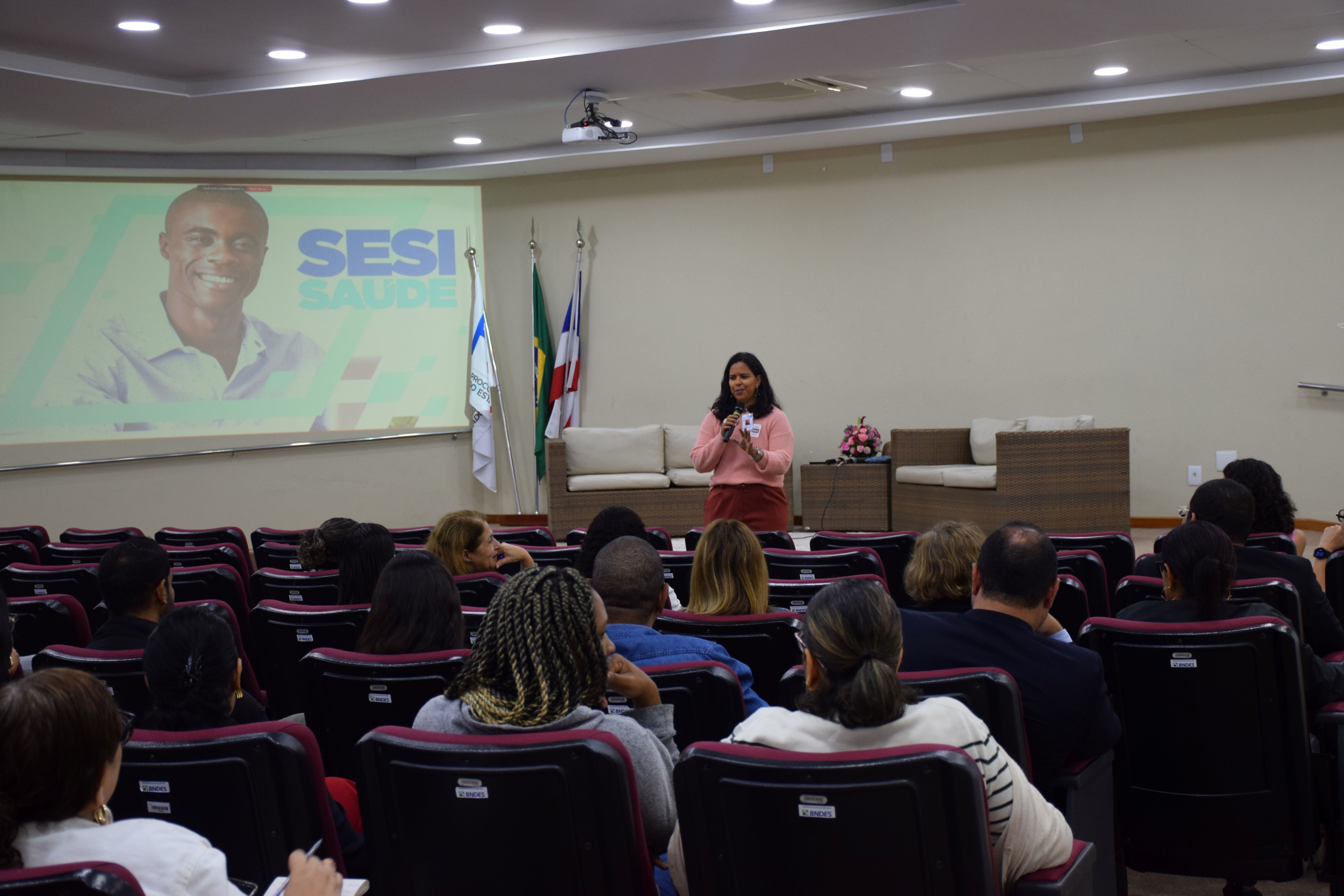 Em um auditório, uma mulher fala ao público segurando um microfone. Ela veste blusa rosa e calça marrom. À frente, há várias fileiras de cadeiras ocupadas por pessoas atentas. No fundo, há um telão com a imagem de um homem sorridente e o texto “SESI SAÚDE”. Próximo ao telão, estão três bandeiras e dois sofás de fibra com almofadas claras. O ambiente é bem iluminado e organizado, com clima de palestra ou apresentação.