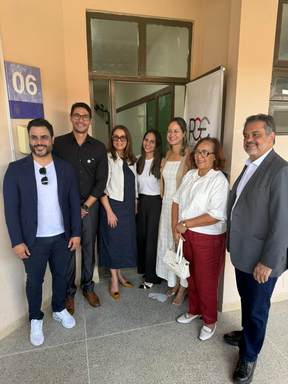 Sete pessoas estão em pé, sorrindo para a câmera, em frente a uma parede bege com uma porta de vidro e uma faixa com o logo "PGE". São quatro mulheres e três homens, vestidos com roupas sociais ou casuais elegantes. À esquerda, um homem de blazer azul e camiseta branca. Ao centro, duas mulheres usam roupas claras. À direita, uma senhora com blusa branca e calça vermelha segura uma bolsa clara, ao lado de um homem de terno cinza. Todos parecem felizes e em um ambiente profissional.
