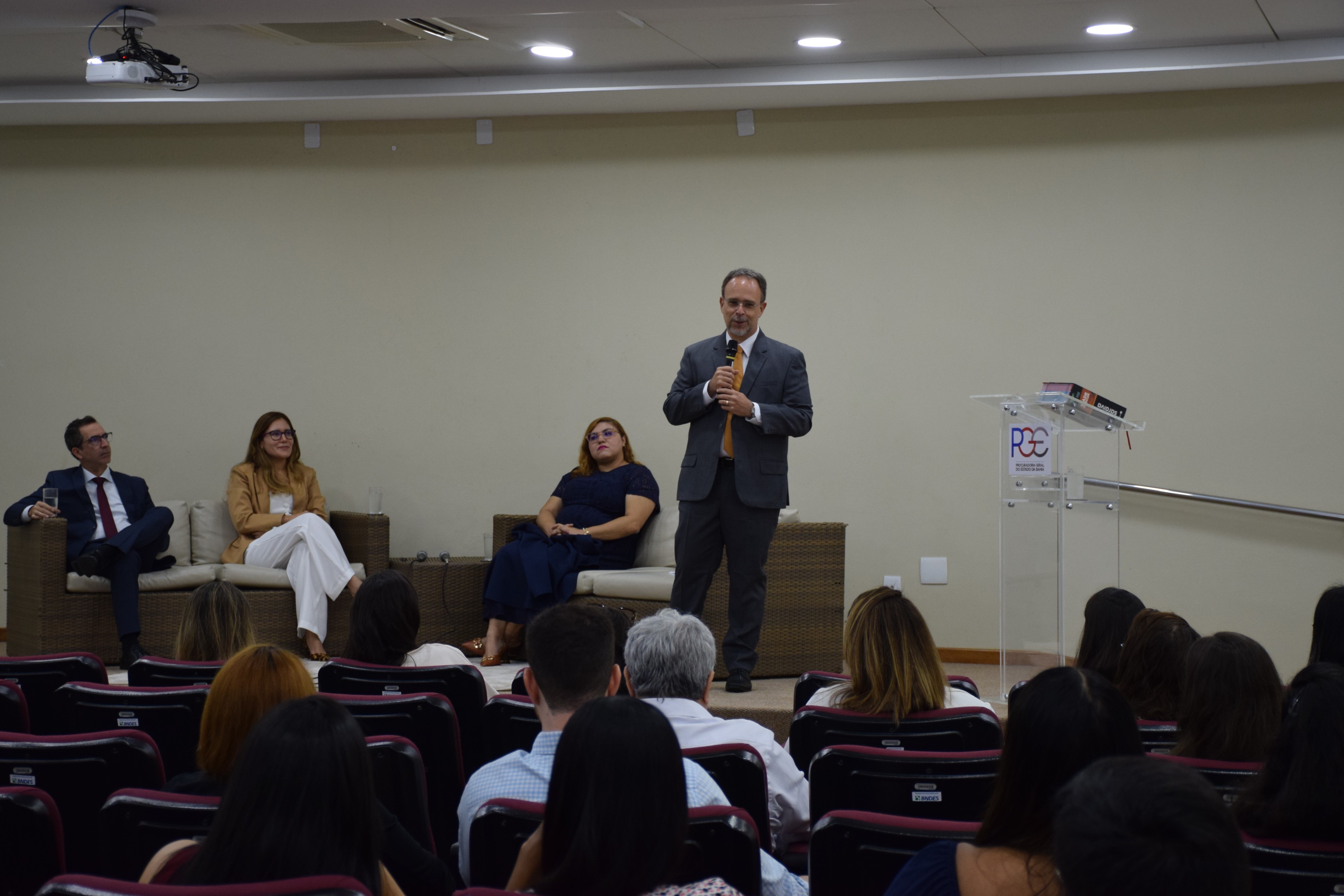 Auditório com várias pessoas sentadas em cadeiras estofadas vinho, voltadas para o palco. No palco, três pessoas estão acomodadas em sofás de fibra: um homem de terno azul, uma mulher de blazer bege e calça branca e outra de vestido azul-marinho. Em pé, à direita, um homem de terno cinza e gravata amarela fala ao microfone próximo a um púlpito transparente com o logotipo da PGE-BA. O fundo é uma parede bege lisa e o ambiente é iluminado por luzes embutidas no teto.
