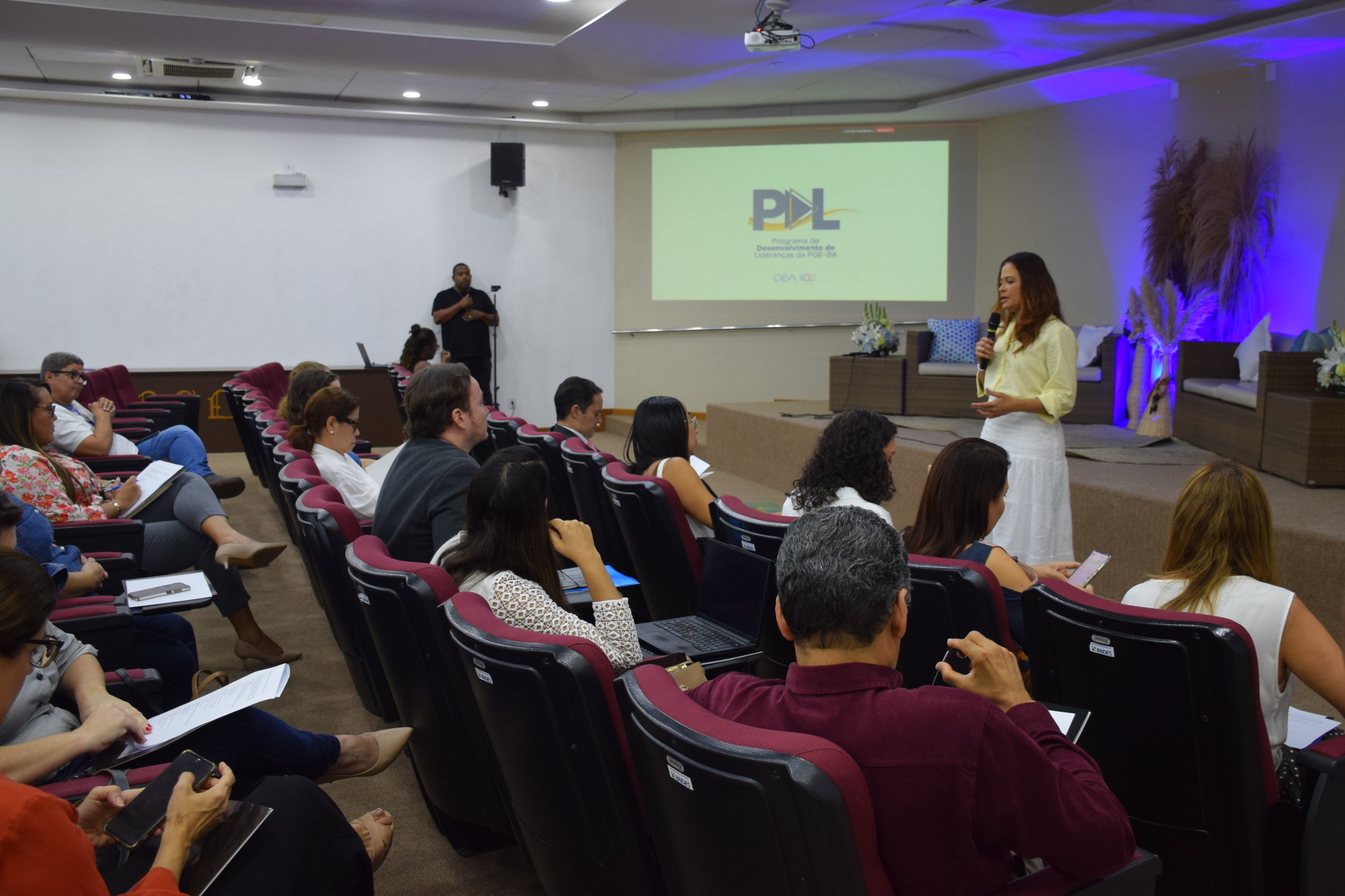 Uma mulher fala em pé diante de um auditório, segurando um microfone. Ela veste blusa amarela clara e saia branca. As pessoas estão sentadas em cadeiras vinho, observando e fazendo anotações. No fundo, há um telão com o logotipo do “Programa de Desenvolvimento de Lideranças da PGE-BA”. Um homem ao lado do auditório faz interpretação em Libras. O ambiente é iluminado e organizado, com clima de evento institucional.