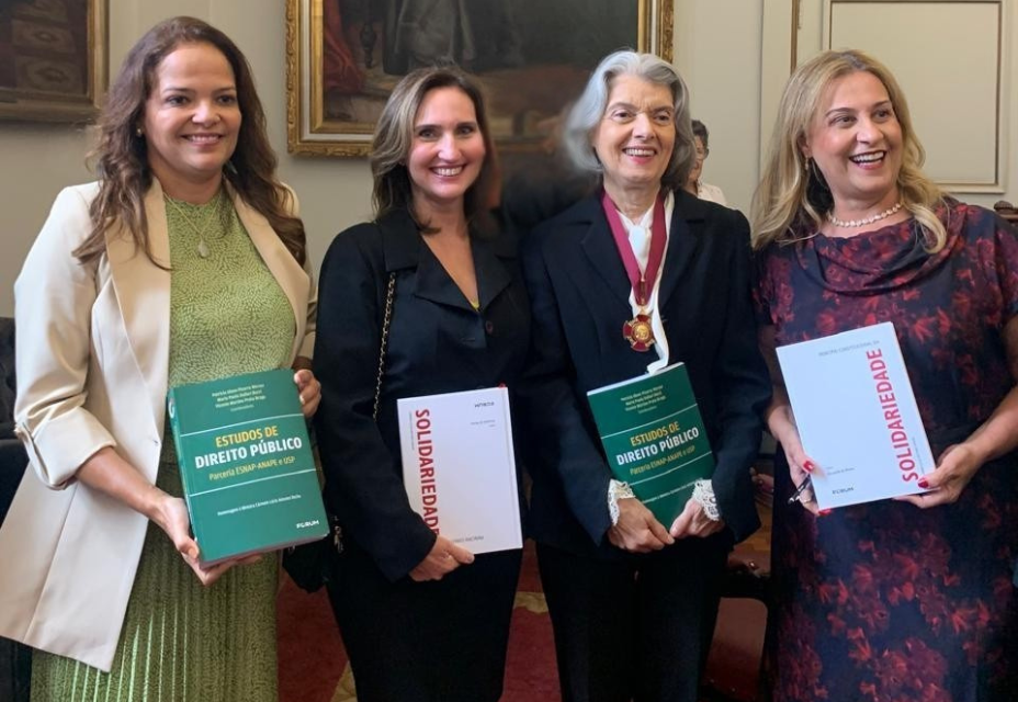 Quatro mulheres estão em pé, sorrindo, lado a lado, em um ambiente formal com quadros nas paredes. Duas delas seguram um livro verde intitulado "Estudos de Direito Público" e as outras duas seguram um livro branco com a palavra "Solidariedade" em vermelho. A terceira mulher, de cabelos grisalhos e roupa preta, usa uma medalha no pescoço. Todas estão bem-vestidas e aparentam estar em um evento jurídico-acadêmico.
