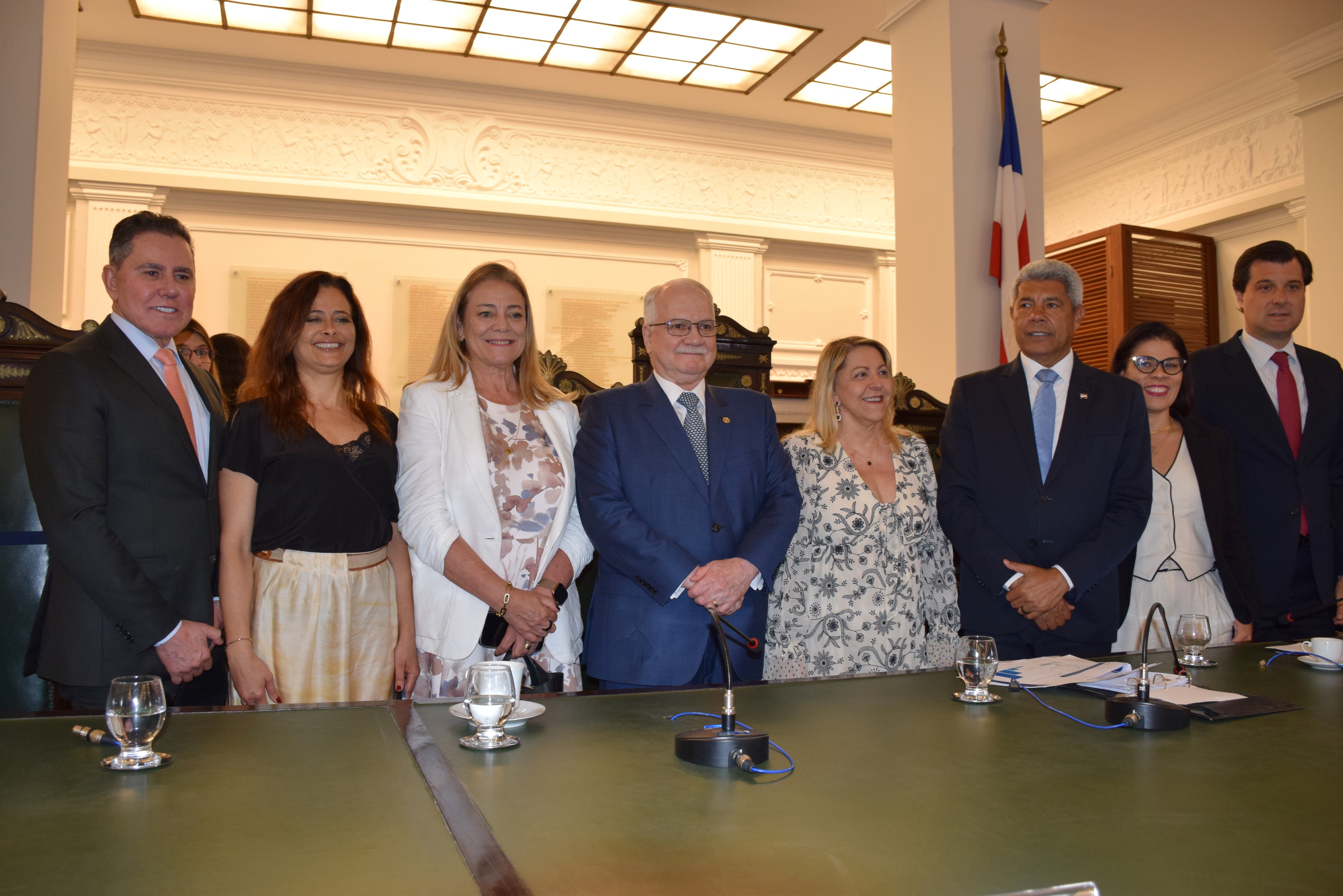 Oito pessoas posam em pé atrás de uma mesa de madeira verde, com microfones e copos d’água sobre ela. Todas estão sorrindo e vestidas de forma formal, com ternos, vestidos e blazers. O ambiente é iluminado e tem bandeiras da Bahia e do Brasil ao fundo, além de detalhes clássicos nas paredes e no teto, indicando um evento institucional.