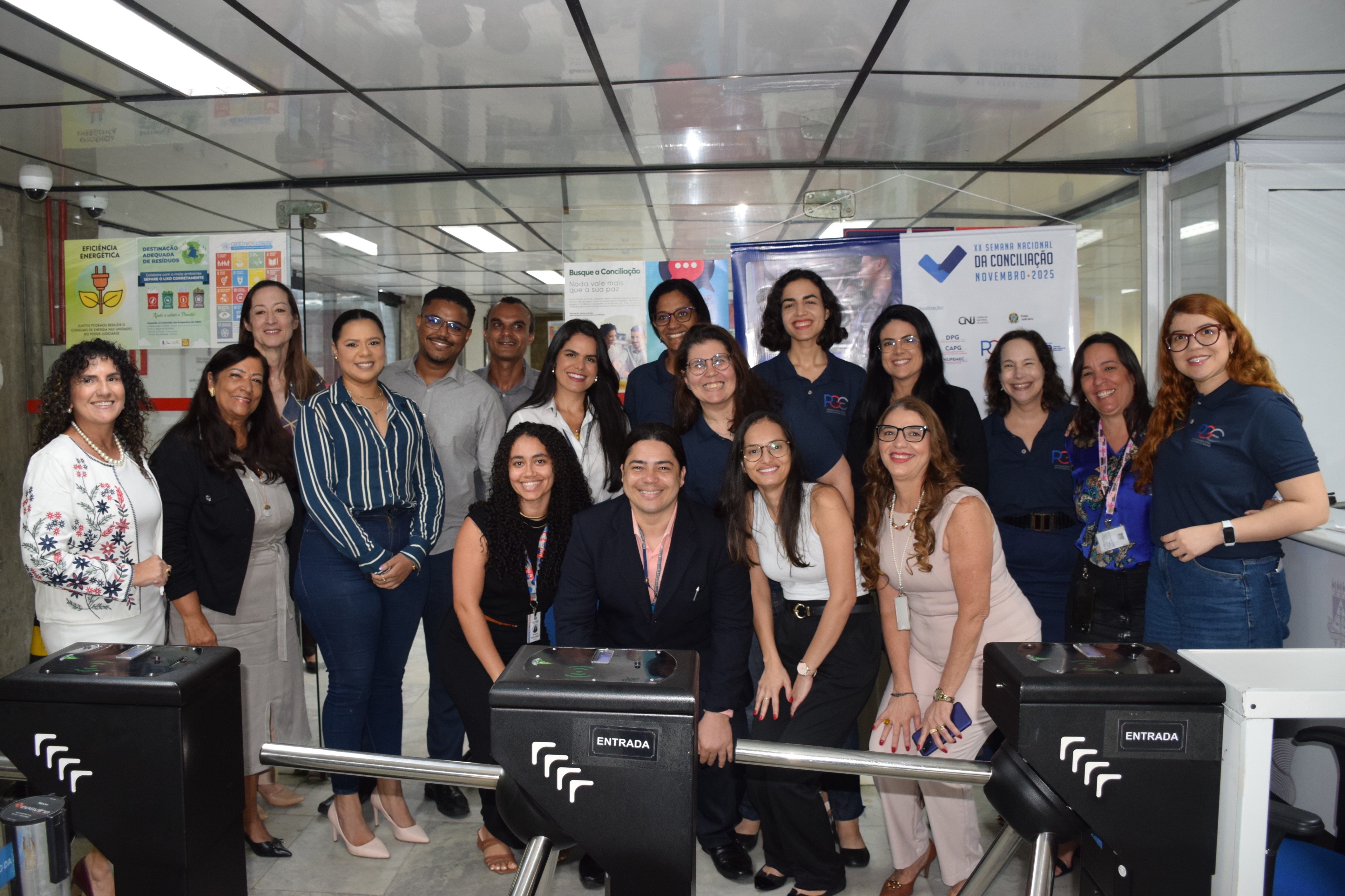 Um grupo de vinte pessoas, composto por homens e mulheres, posa sorrindo para a câmera em um ambiente interno. A maioria veste roupas sociais ou casuais escuras. O grupo está de pé, exceto por um homem no centro, agachado, e uma mulher à sua frente. Eles se agrupam próximos a duas catracas pretas, com a palavra "ENTRADA" em branco. Ao fundo, painéis e cartazes coloridos estão em exposição. As luzes de teto são fluorescentes.