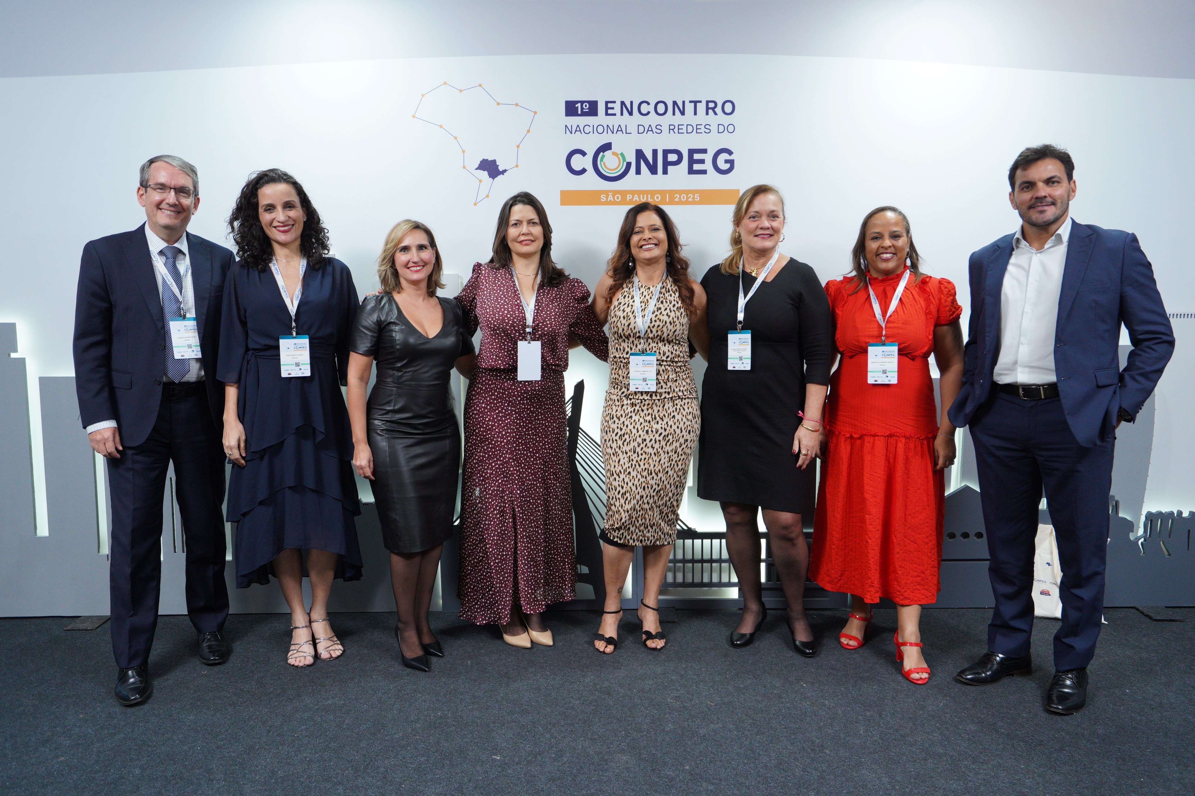  A imagem mostra oito pessoas posando para foto em frente a um painel de evento. Elas estão alinhadas lado a lado, sorrindo, e usam roupas sociais e crachás. Há homens e mulheres no grupo, todos em postura formal e amistosa. O painel atrás é claro, com logotipos repetidos. O ambiente parece ser de congresso ou encontro profissional.