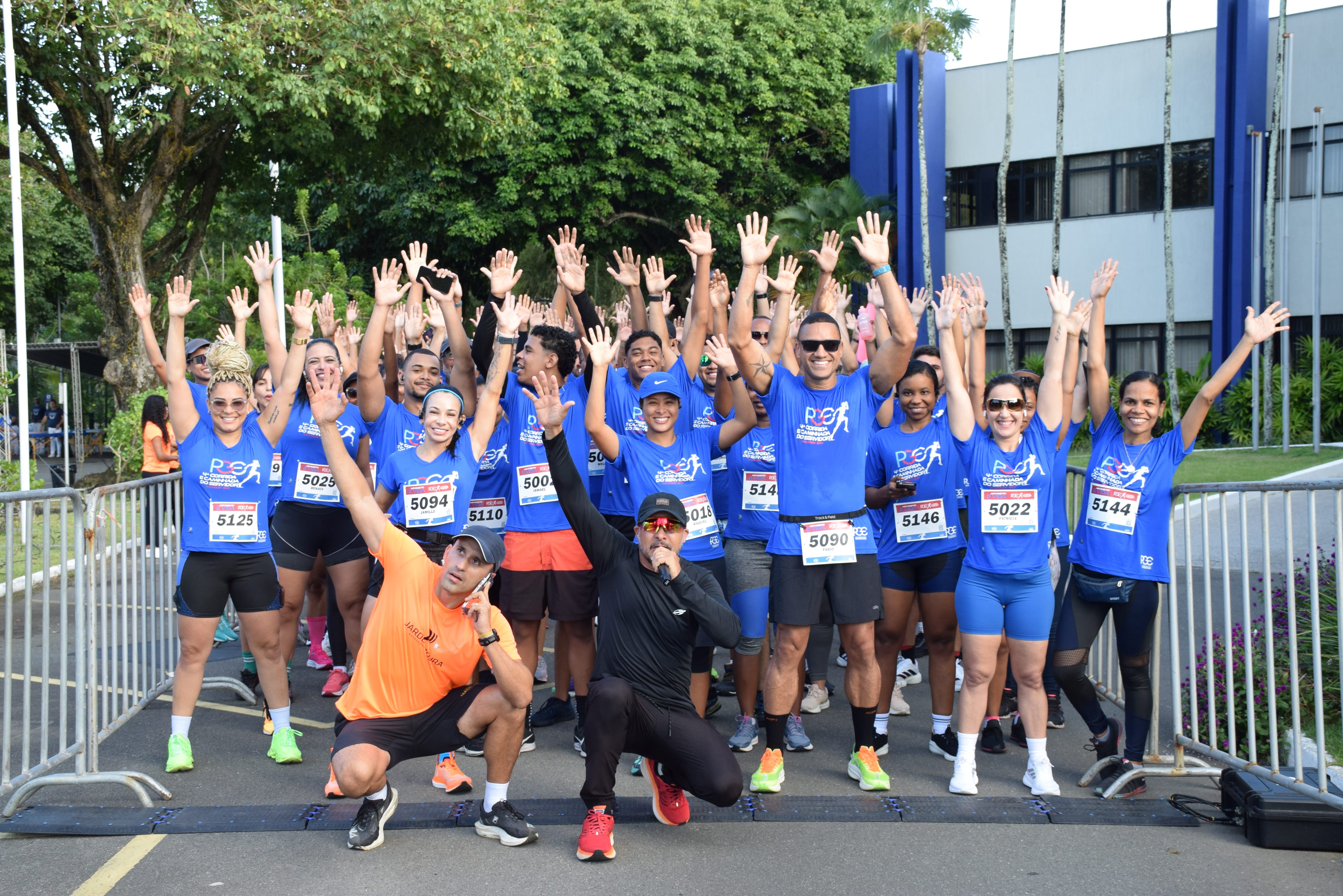 Foto que mostra um grupo de cerca de 20 corredores de rua, sorridentes, de camiseta azul com o logo da corrida. Eles estão em pé, com os braços levantados, logo após a linha de chegada de uma corrida. Dois homens estão agachados na frente.