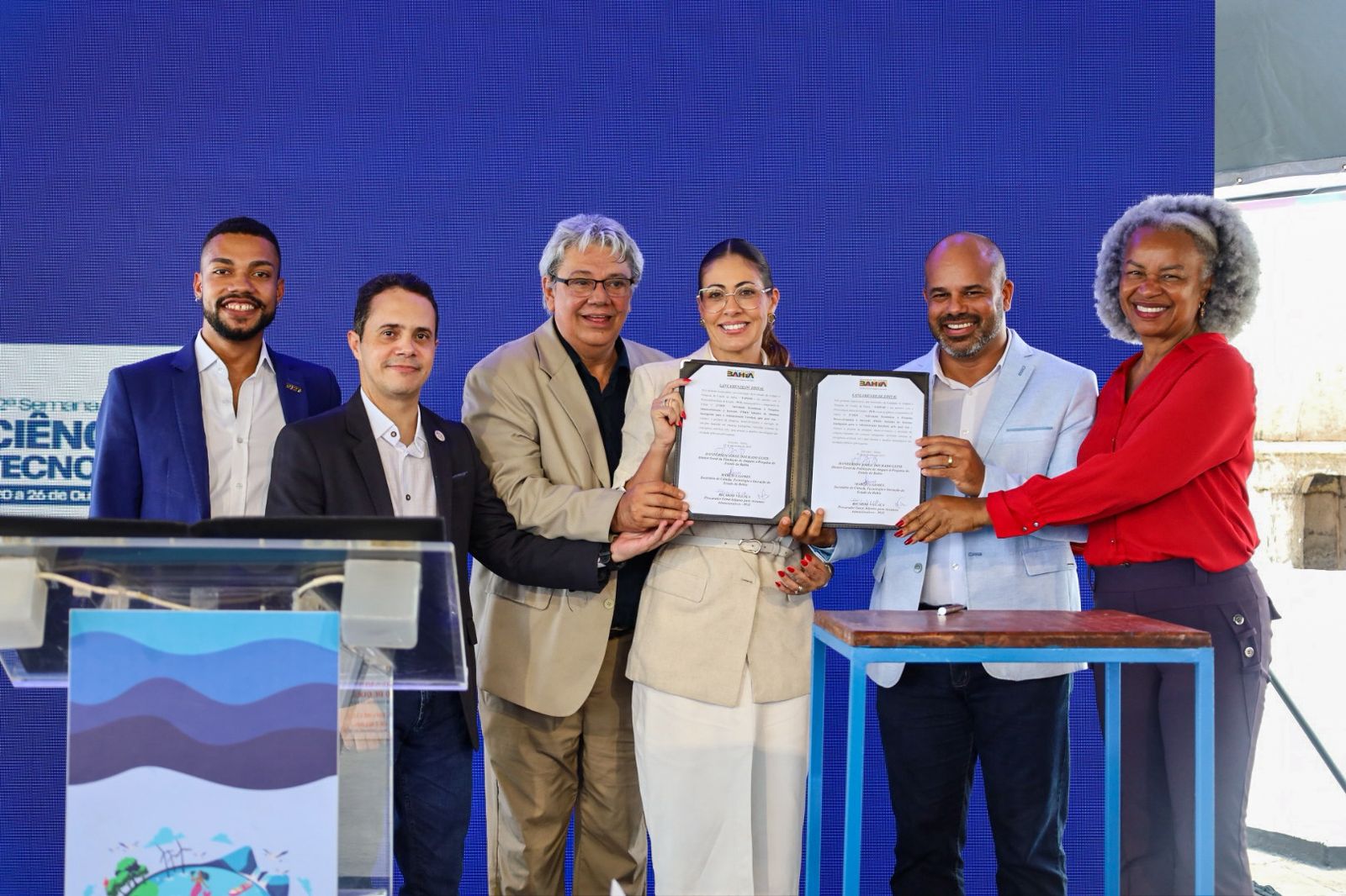 Seis pessoas, quatro homens e duas mulheres, estão em pé em frente a um painel azul. O grupo segura um documento em conjunto.  Da esquerda para a direita: um homem negro de blazer azul; um homem branco de camisa social preta; um homem branco de blazer bege; uma mulher branca de blazer e calça bege claro; um homem negro de blazer azul claro; e uma mulher negra de camisa vermelha. Todos sorriem.