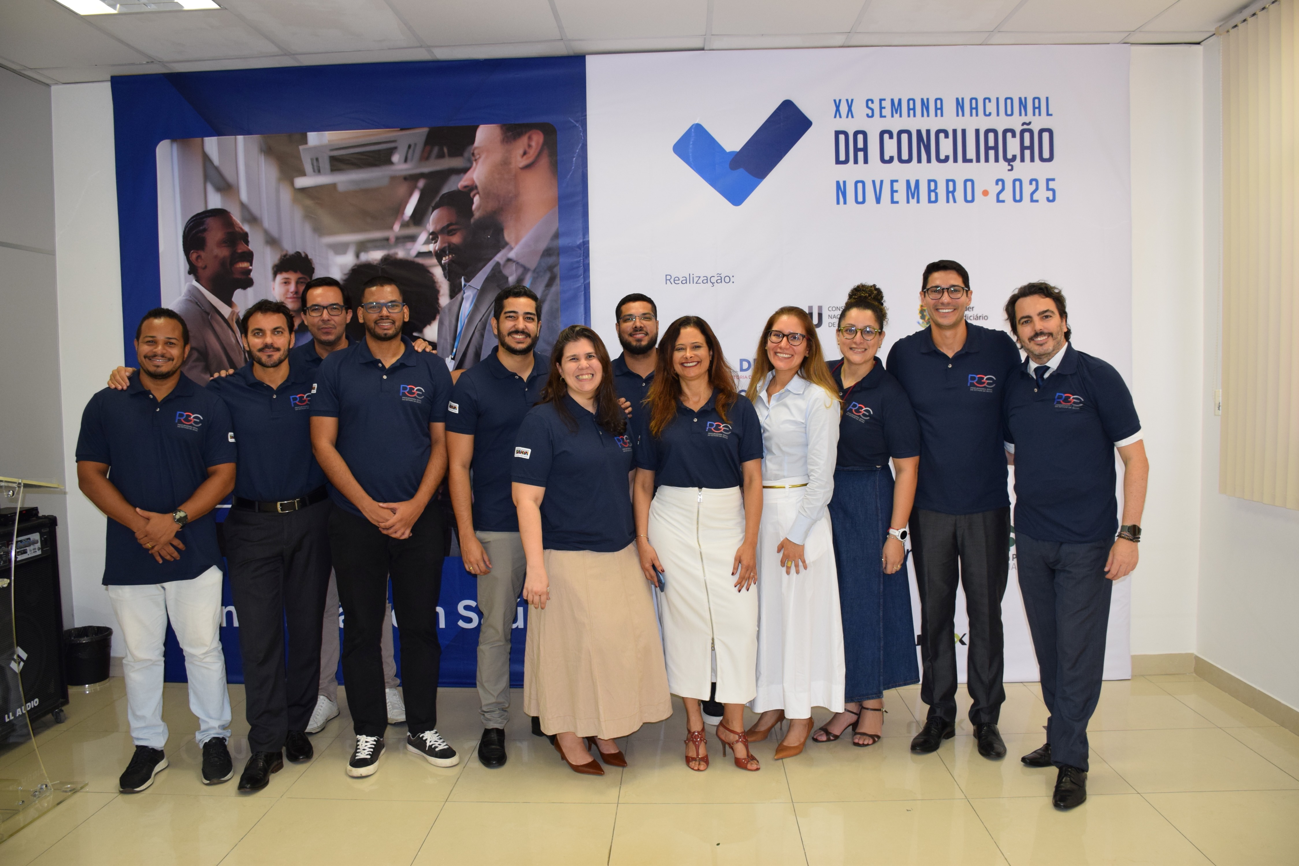 Um grupo de doze pessoas posa sorridente em um ambiente interno, diante de um grande banner azul e branco com o texto “XX Semana Nacional da Conciliação – Novembro 2025”. A maioria veste camisa polo azul-marinho com o logotipo da PGE, calças sociais ou saias em tons neutros. O grupo está lado a lado, demonstrando integração e alegria. O local tem piso claro, parede branca e cortina bege à direita.