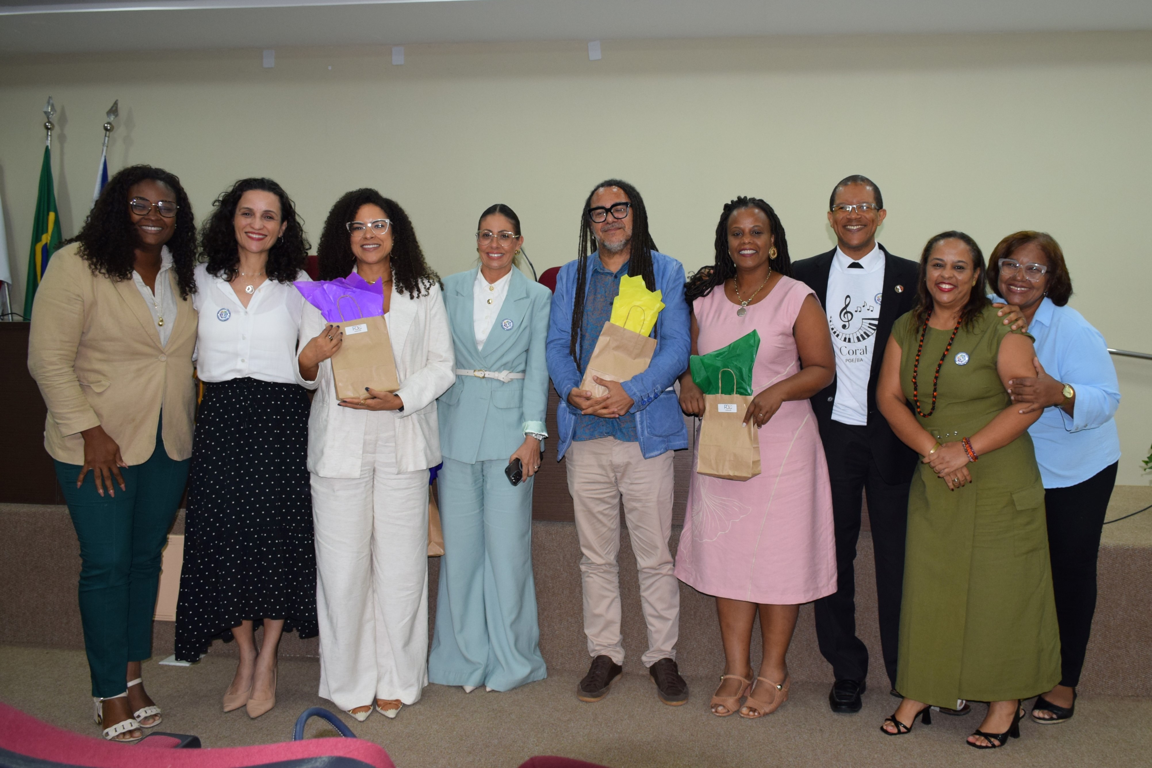Oito pessoas posam sorridentes em frente ao palco de um auditório. O grupo está bem vestido, com roupas sociais e elegantes, e alguns seguram saquinhos de presente coloridos. Ao fundo, há bandeiras e um púlpito com o logotipo da PGE. O clima é de confraternização e encerramento de um evento.