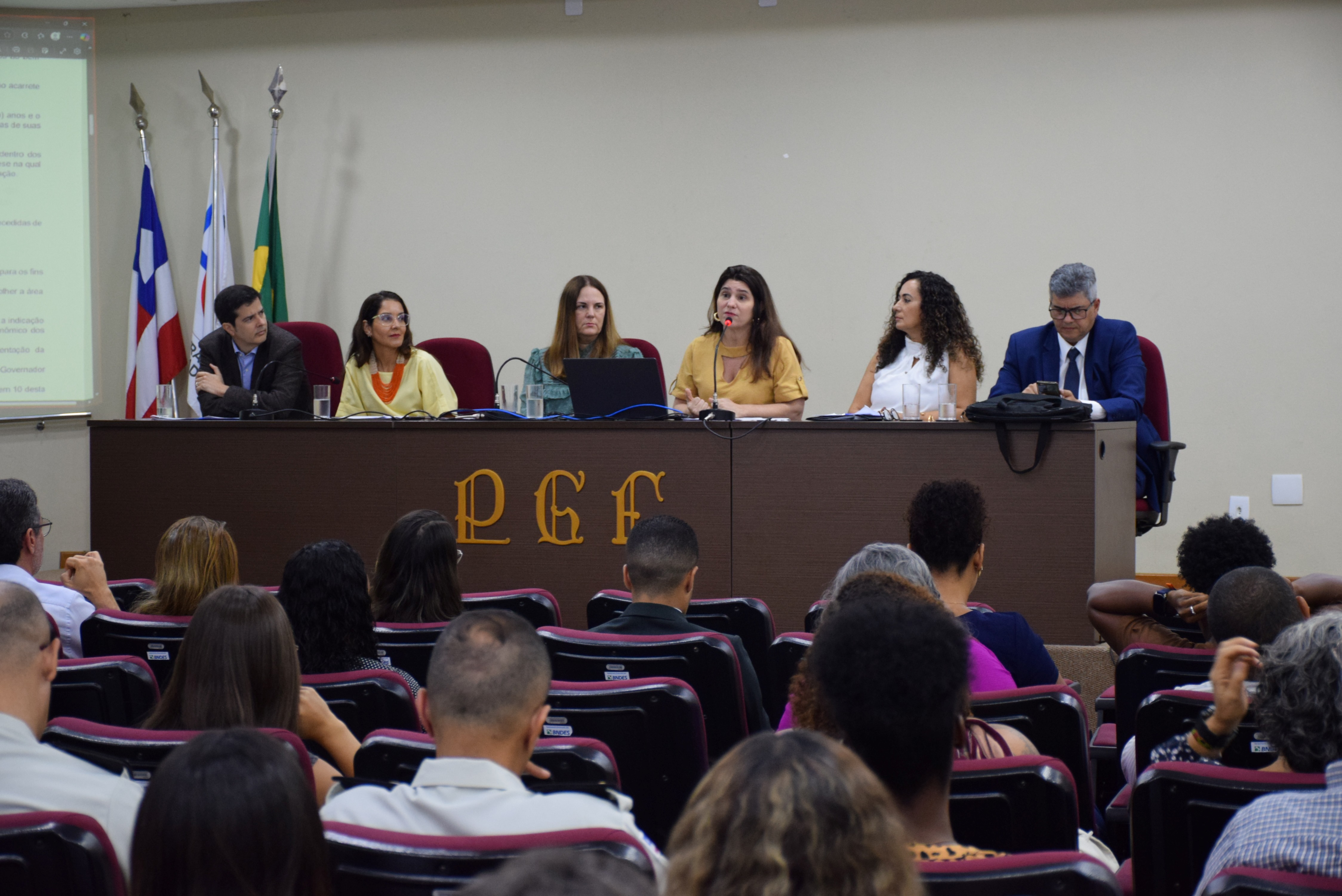 A imagem mostra seis pessoas sentadas lado a lado em uma mesa longa, participando de um evento formal. Há quatro mulheres e dois homens, todos vestidos de forma elegante. Atrás deles, estão três bandeiras: uma do Brasil, uma estadual e a terceira da PGE-BA. À frente da mesa, está a sigla "PGE" em letras grandes. Na parte inferior, algumas pessoas assistem à apresentação. O ambiente é uma sala de conferências com iluminação clara e um telão ao lado esquerdo. A cena transmite uma reunião oficial.