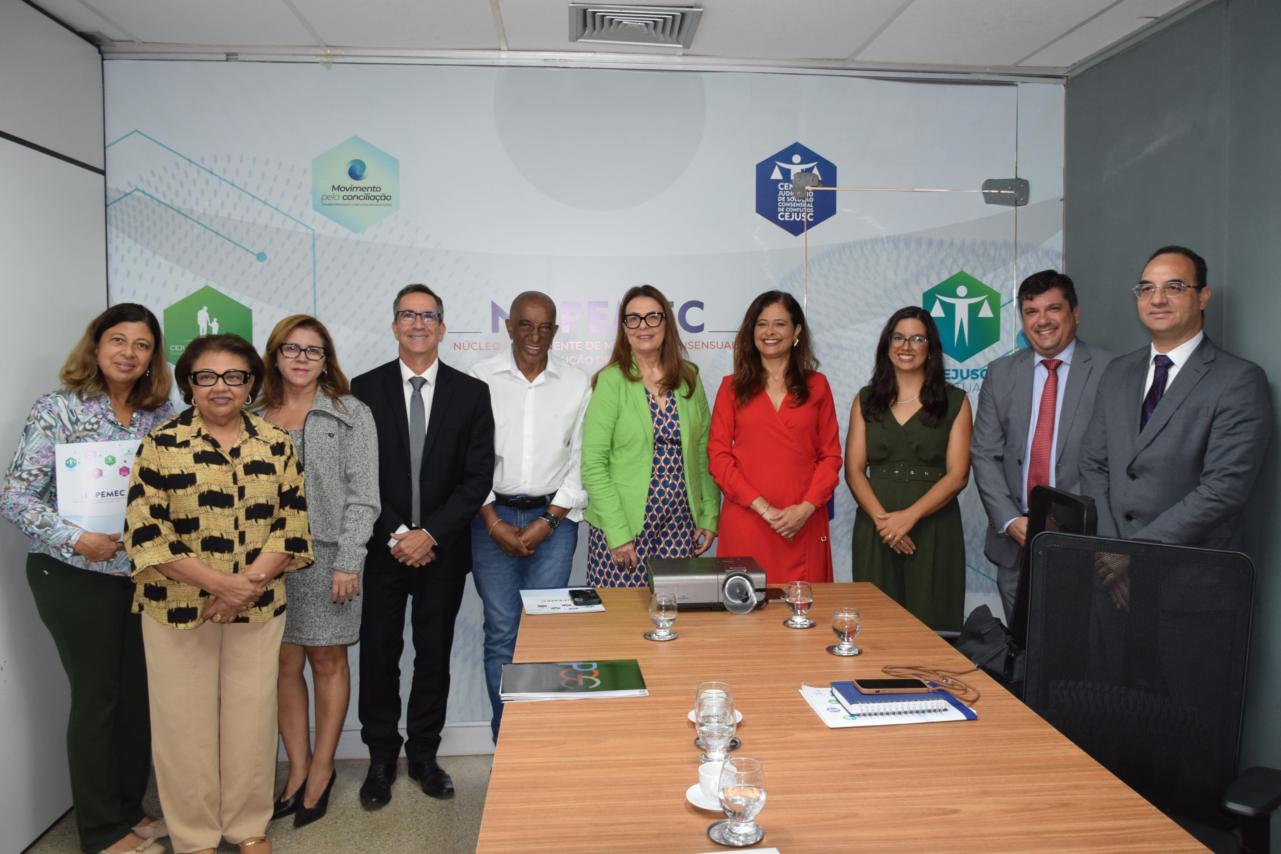 Grupo de oito pessoas em pé, dentro de uma sala de reuniões, conversando em frente a uma parede com logotipos do Poder Judiciário e do CEJUSC.