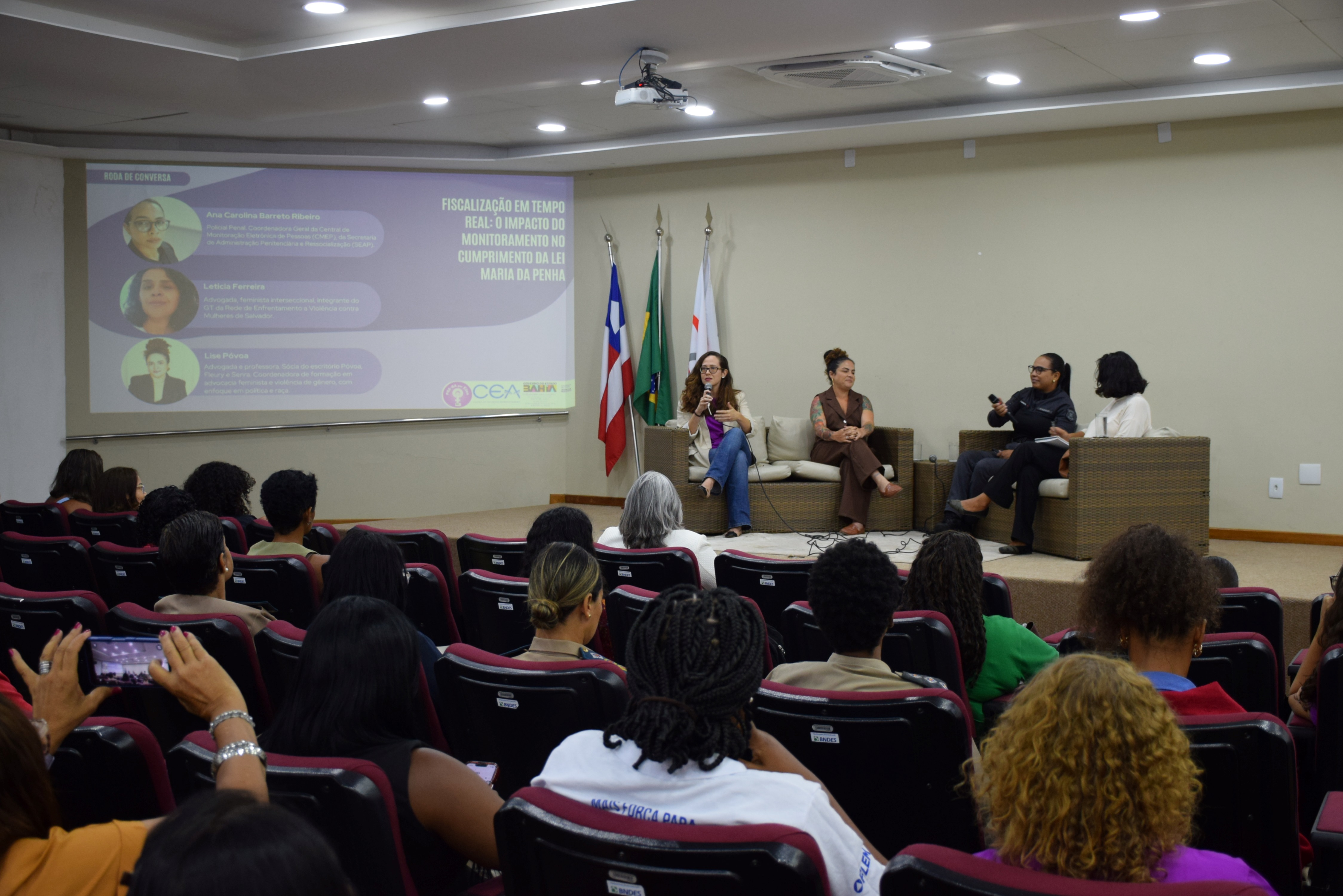Em um auditório, várias pessoas estão sentadas em fileiras de cadeiras, formando o público e voltadas para o palco. No palco, quatro mulheres adultas estão sentadas em sofás claros, participando de uma roda de conversa. Uma delas segura o microfone e fala, enquanto as outras escutam. Ao fundo, um telão exibe informações do evento sobre fiscalização em tempo real e a Lei Maria da Penha. À direita do palco, estão as bandeiras da Procuradoria Geral, de Salvador e da Bahia. 