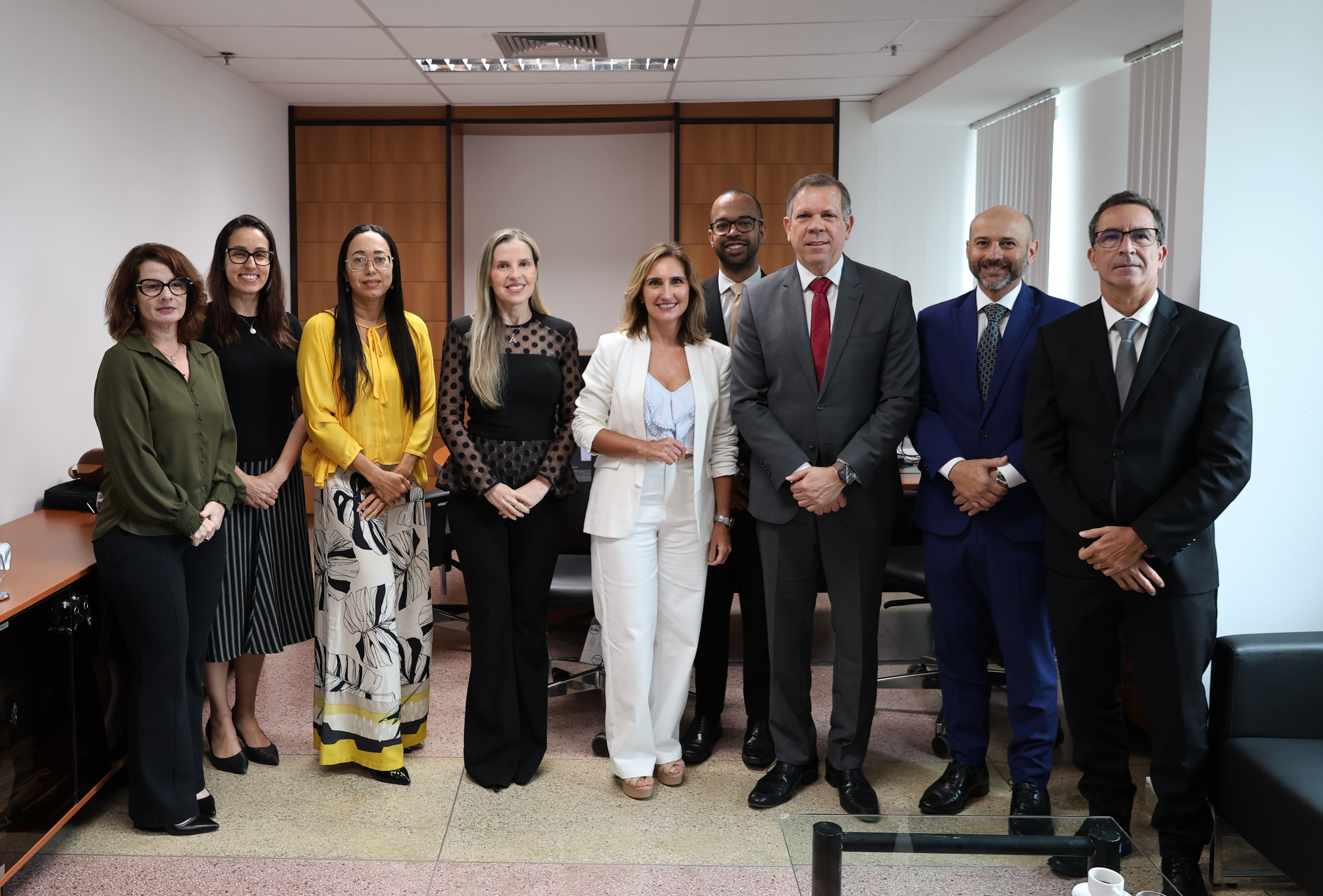 Nove pessoas estão alinhadas, posando para uma foto formal em um ambiente de escritório. São cinco mulheres e quatro homens, todos vestidos com roupas sociais. As mulheres usam blusas e calças ou saias em tons variados, incluindo amarelo, preto e branco. Os homens vestem ternos escuros com gravatas coloridas. O ambiente é claro, com paredes brancas, janelas com persianas e móveis de escritório ao fundo. Todos estão de pé, sorrindo ou com expressão séria, transmitindo profissionalismo.