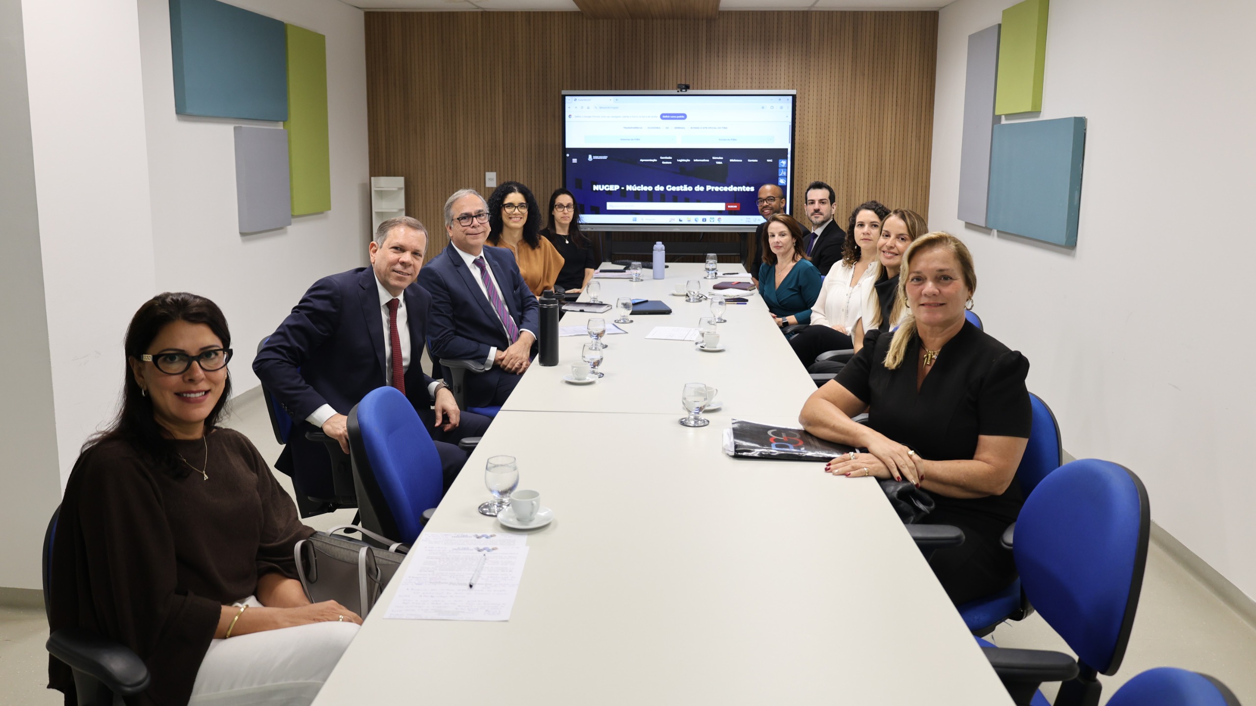Sala de reunião com mesa longa e pessoas sentadas ao redor. Homens e mulheres, em trajes formais, participam de uma conversa. Alguns anotam, outros escutam com atenção. Ao fundo, uma tela exibe o título “Semana de Saneamento, Precedentes e Ações Coletivas”. O ambiente é claro, com paredes neutras e painéis coloridos.