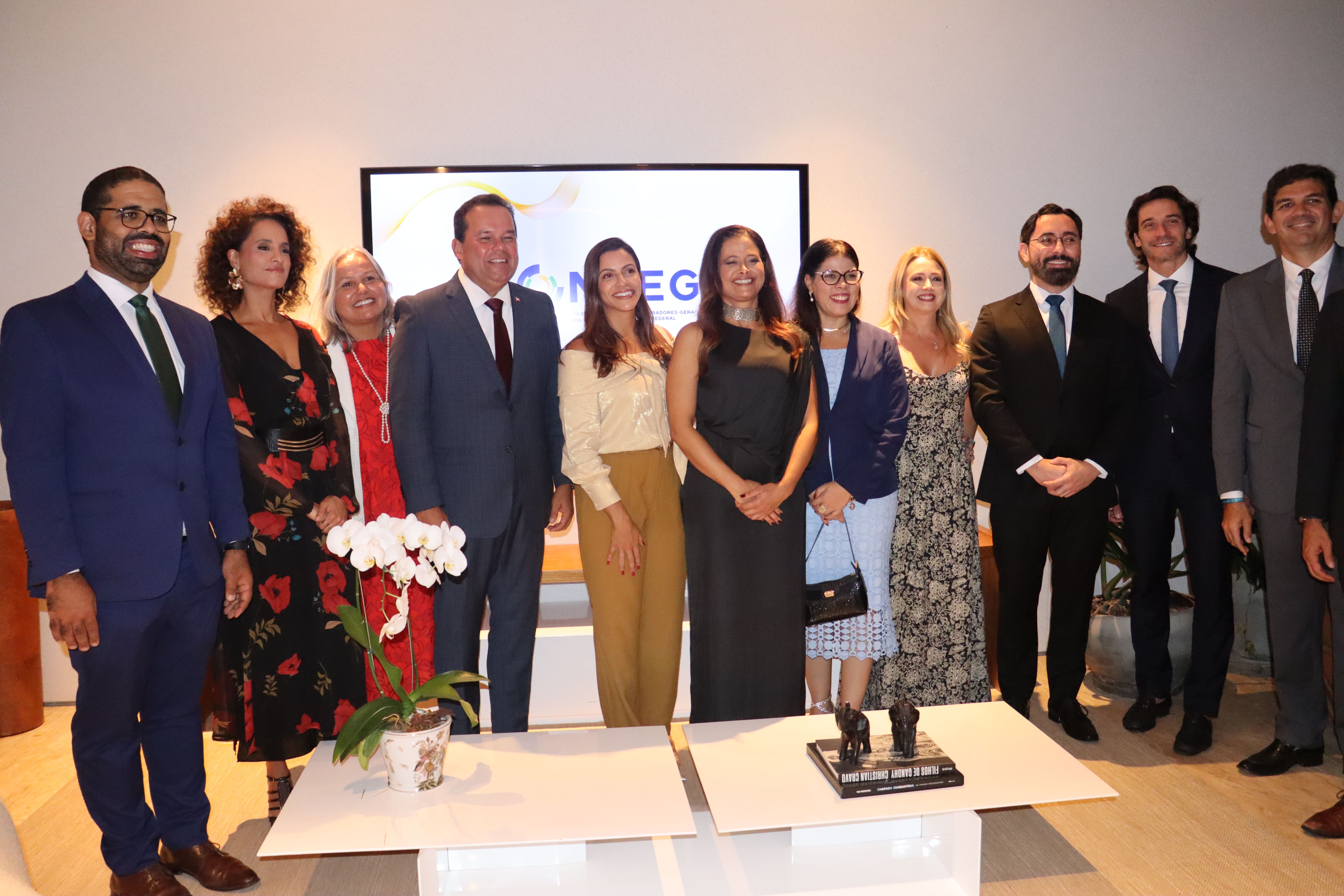 Grupo de várias pessoas alinhadas para uma foto em evento formal. Homens e mulheres vestem roupas sociais e estão sorrindo. Ao fundo, há um telão com logotipo e decoração simples, indicando uma cerimônia ou encontro institucional.