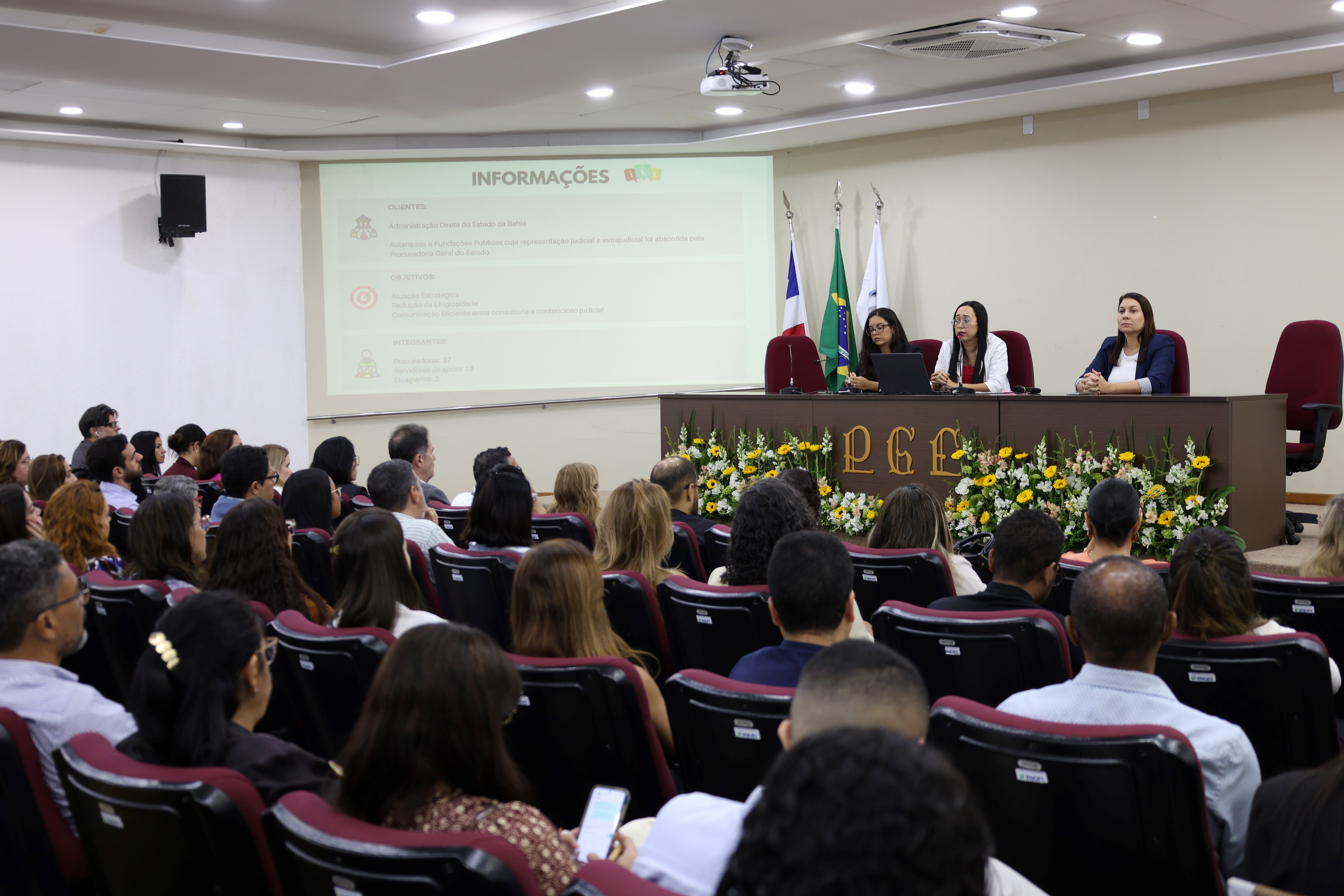 Foto de um auditório durante uma palestra ou evento institucional. Diversas pessoas estão sentadas em fileiras de cadeiras, voltadas para a frente da sala. No palco, três mulheres compõem uma mesa de apresentação. Ao fundo, há uma tela de projeção exibindo informações em formato de slides e bandeiras posicionadas ao lado da mesa. A frente do palco é decorada com arranjos de flores. O ambiente transmite um momento de capacitação, apresentação de conteúdo e troca de conhecimentos entre os participantes.